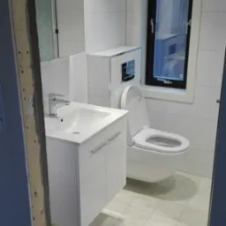 Nowoczesna, jasna łazienka z białą ceramiką, umywalką z szafką, podwieszaną toaletą i oknem z czarną ramą, widok na niedokończone futryny drzwi.