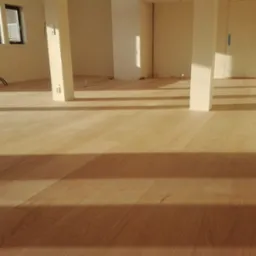 Jasne panele podłogowe w nowym, pustym pomieszczeniu z białymi ścianami i kolumnami, widoczne kable elektryczne przed montażem gniazdek.