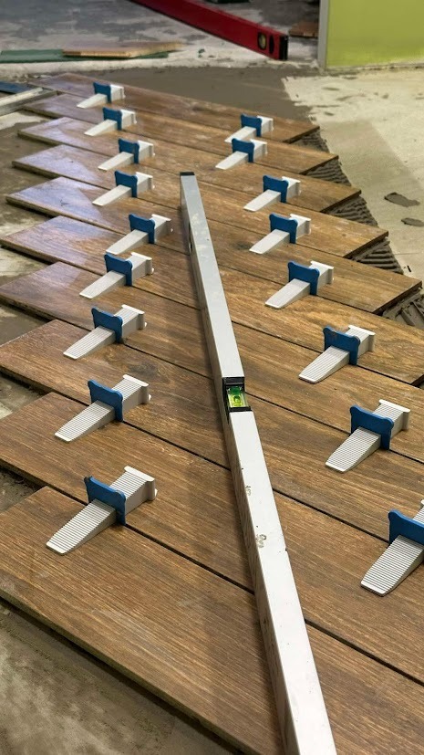 Układanie płytek imitujących drewno z użyciem poziomicy i klipsów poziomujących, widoczne ślady zaprawy.