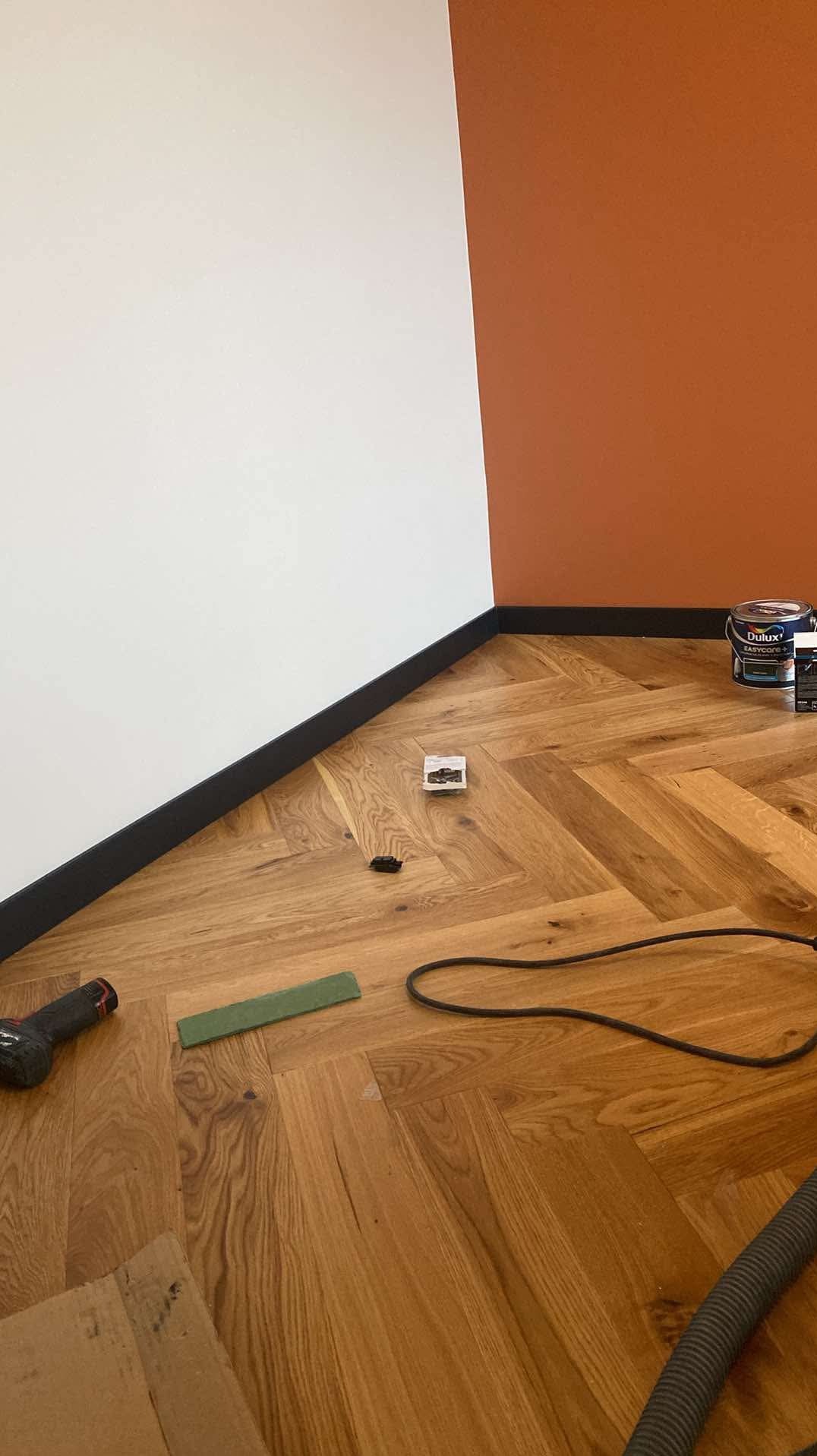 Układanie parkietu w jodełkę w pokoju z pomalowanymi ścianami na biało i terakotowo, widoczne narzędzia, puszka farby Dulux, listwy przypodłogowe w kolorze czarnym.