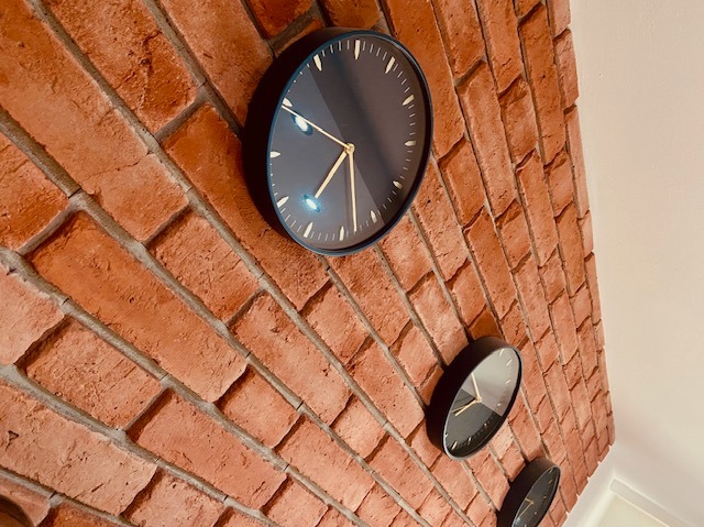 Ceglana ściana z trzema czarnymi zegarami z złotymi wskazówkami, ułożonymi w pionie, w pomieszczeniu z białym sufitem.