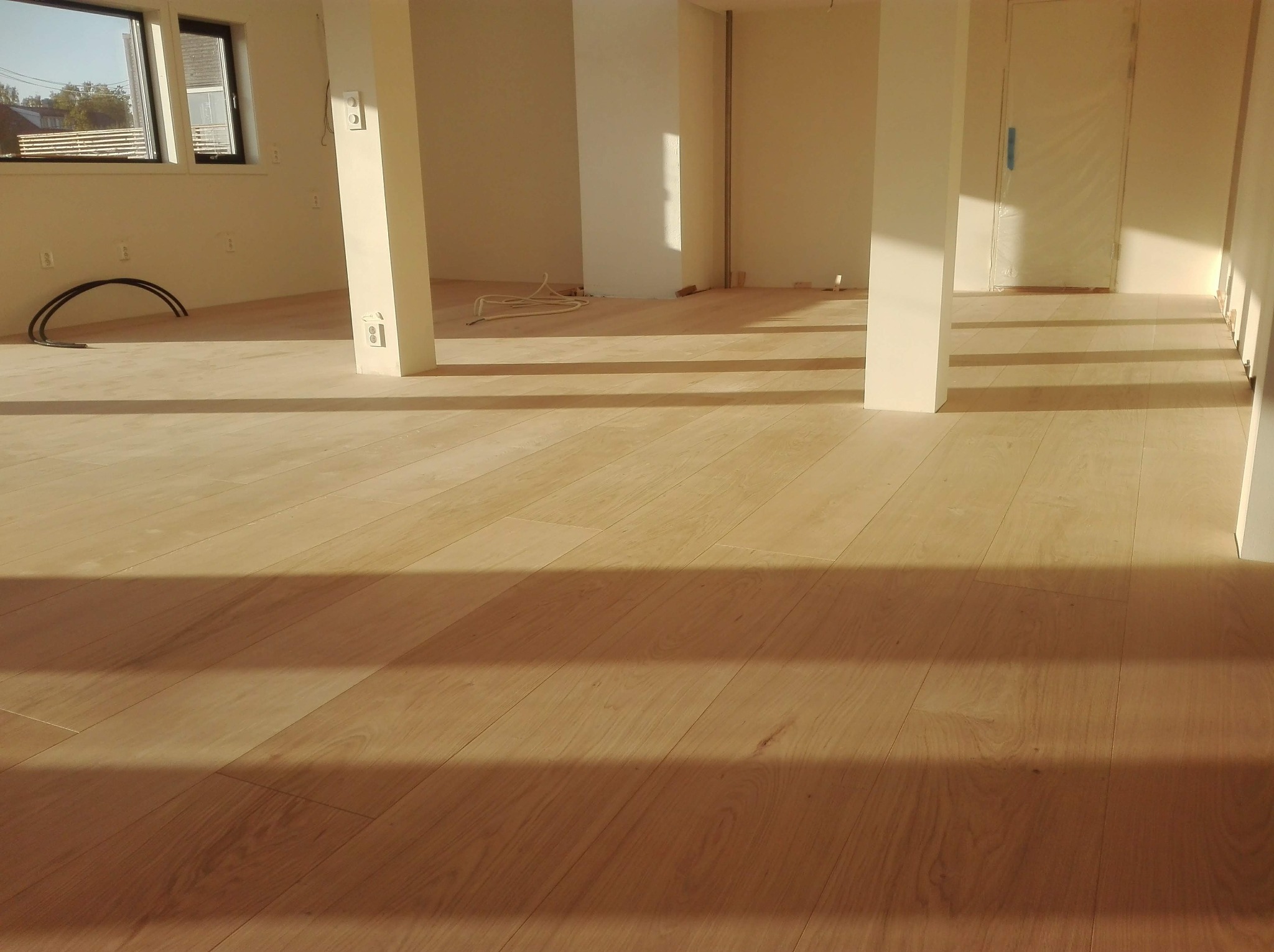 Jasne panele podłogowe w nowym, pustym pomieszczeniu z białymi ścianami i kolumnami, widoczne kable elektryczne przed montażem gniazdek.