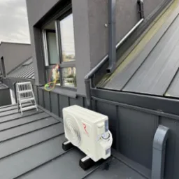 Biały klimatyzator Rotenso na szarym, blaszanym dachu nowoczesnego domu. Widoczna drabina, okno i orynnowanie. Montaż klimatyzacji na dachu.