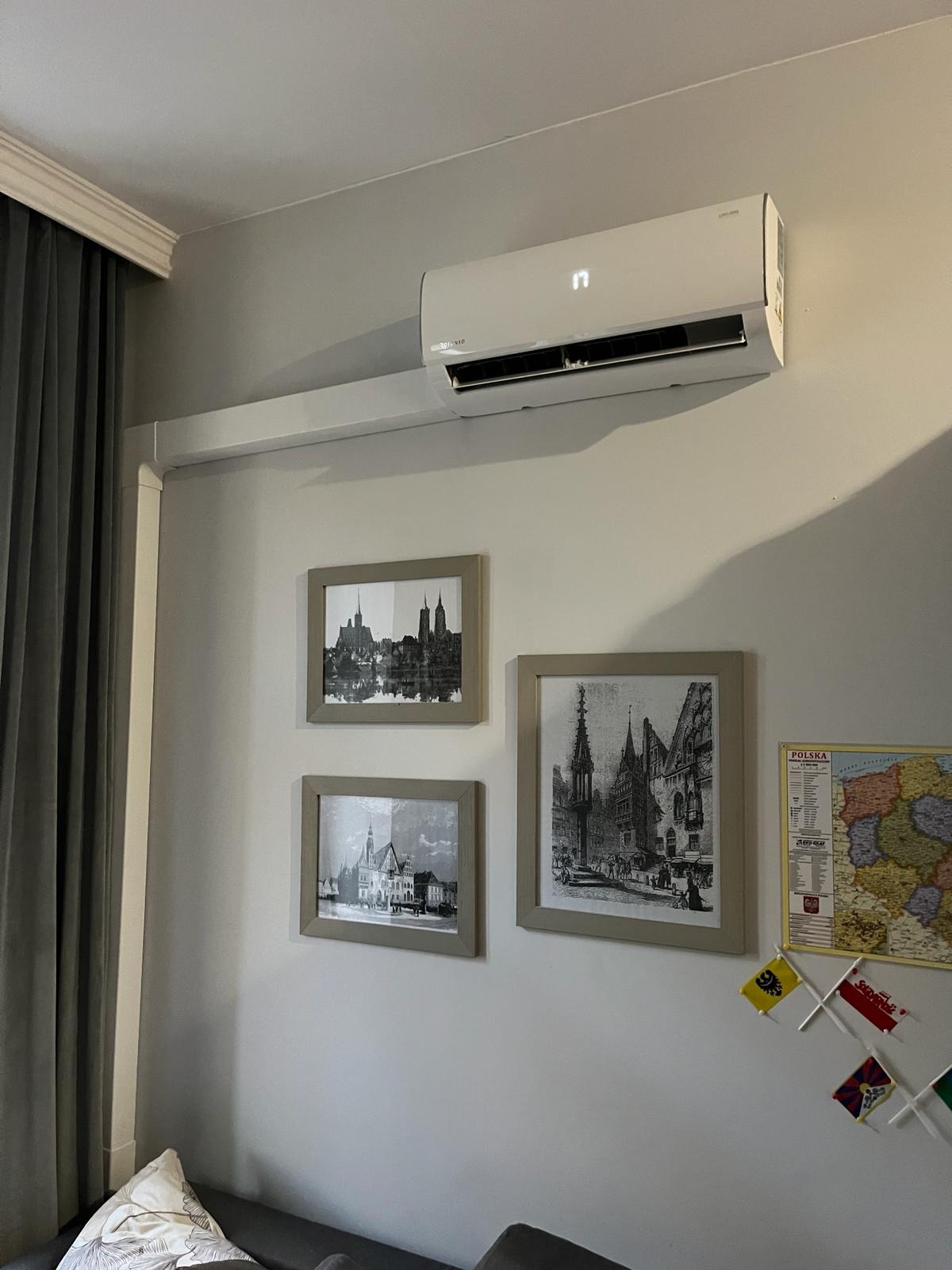 Biała klimatyzacja na ścianie w pokoju, pod nią trzy obrazy w ramkach i mapa Polski. Widoczny fragment zasłony i sofy. Urządzenie włączone, wyświetla temperaturę.