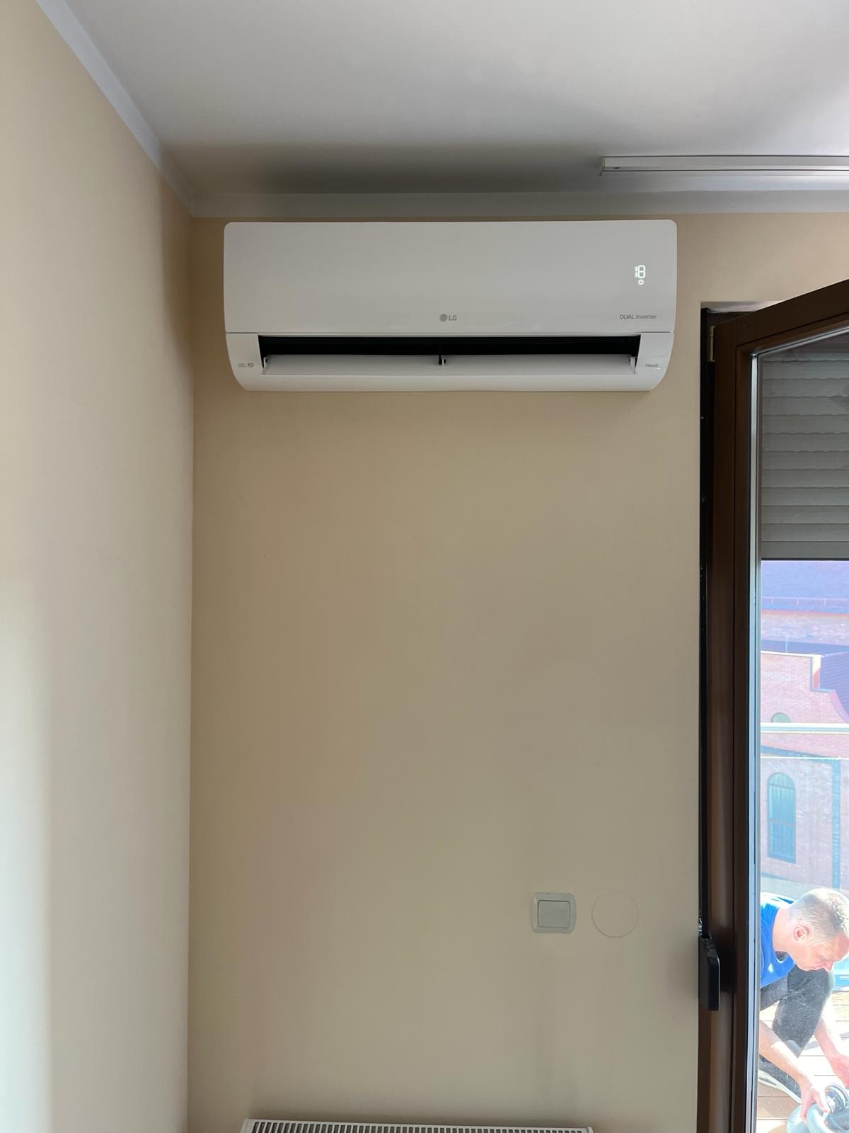 Biały klimatyzator LG Dual Inverter na beżowej ścianie w jasnym pomieszczeniu. Widoczne okno i fragment osoby w tle, temperatura na wyświetlaczu klimatyzacji to 18 stopni.