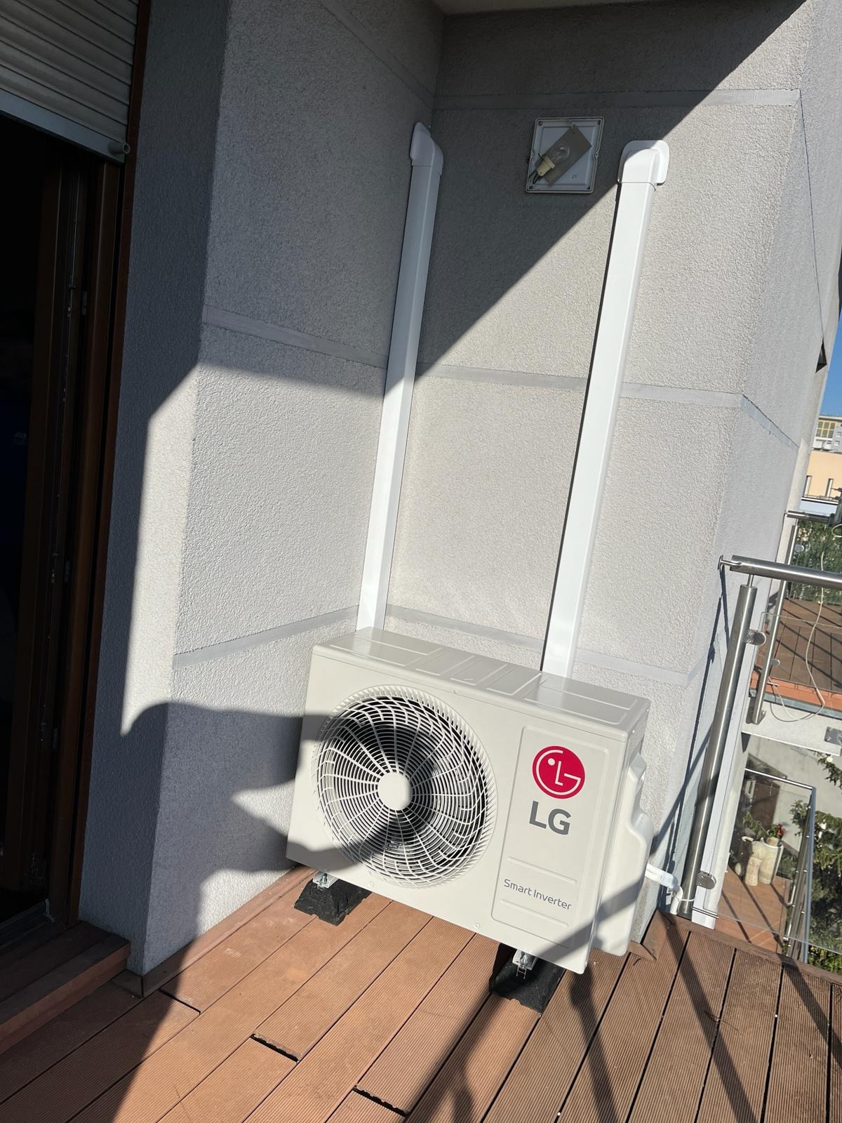 Biała jednostka zewnętrzna klimatyzacji LG Smart Inverter zamontowana na balkonie, z widocznymi rurami odprowadzającymi. Ściana budynku oświetlona słońcem.