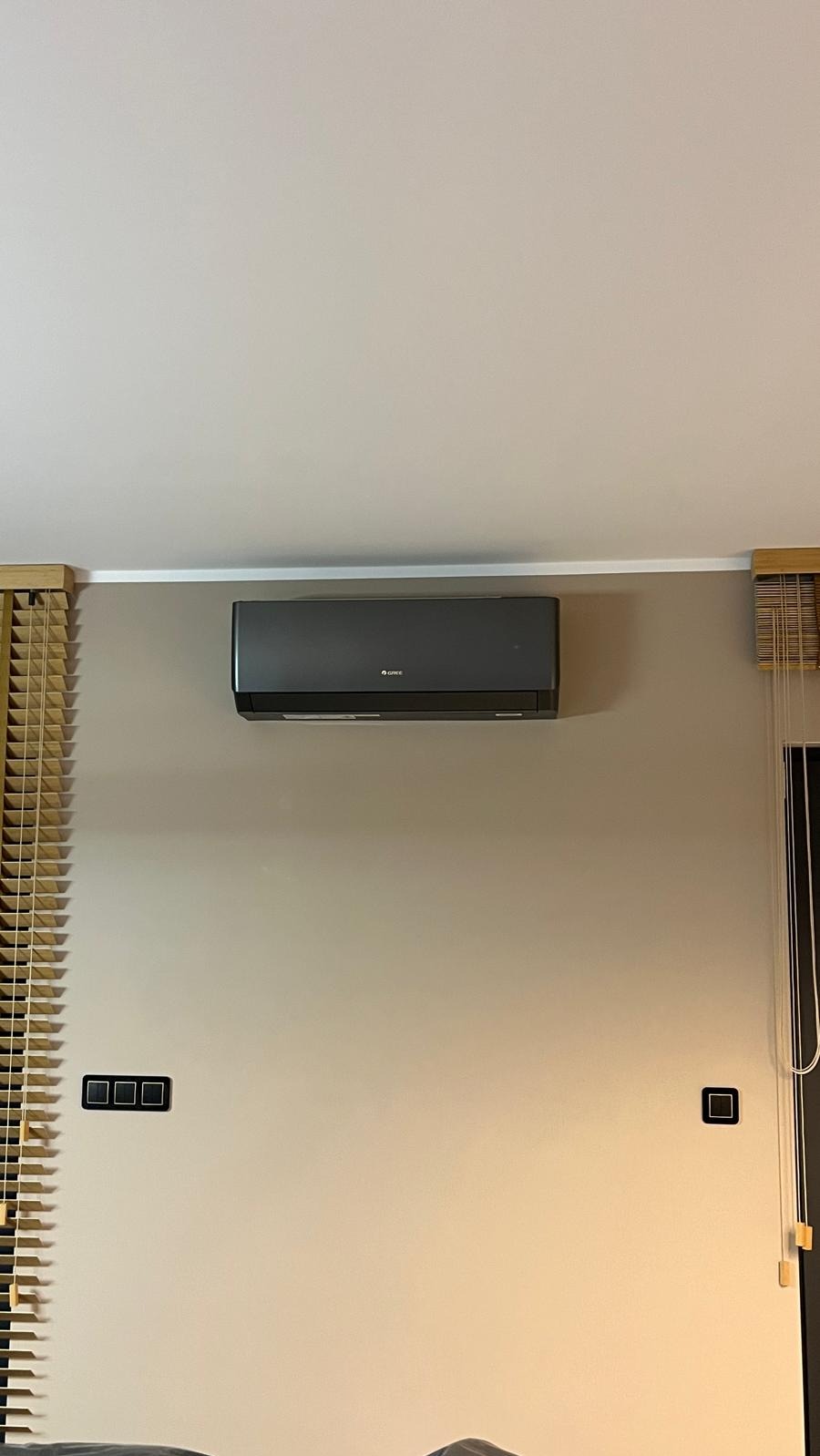Zamontowany, grafitowy klimatyzator Gree na beżowej ścianie z drewnianymi żaluzjami. Widoczne włączniki. Minimalistyczny design wnętrza.