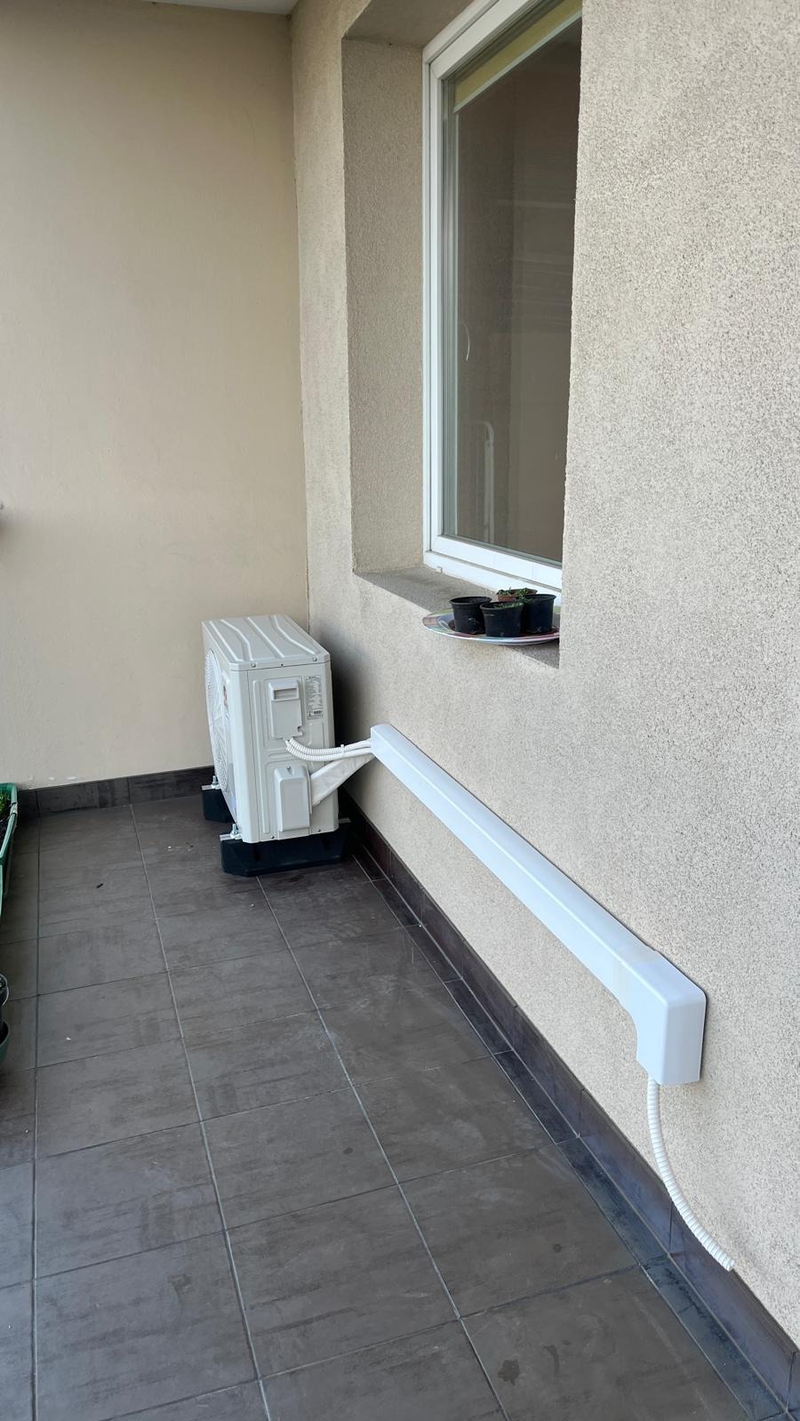 Zewnętrzna jednostka klimatyzacji na balkonie, z białym korytem maskującym przewody, obok okna z doniczkami. Widok z boku, na szarych płytkach.
