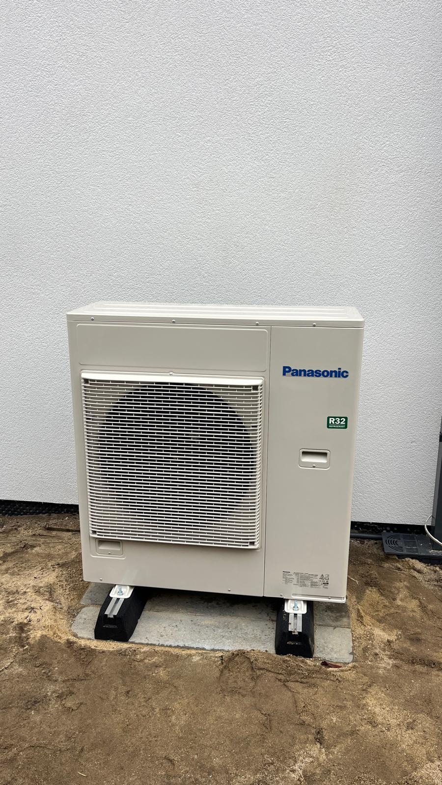 Kompaktowa, beżowa jednostka zewnętrzna klimatyzacji Panasonic R32, zamontowana na betonowej podstawie z podkładkami antywibracyjnymi, na tle białej ściany.