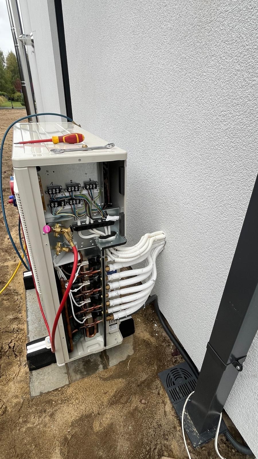 Otwarta obudowa klimatyzatora zewnętrznego z widocznymi przewodami, instalacją elektryczną i narzędziami na górze, umieszczona na zewnątrz budynku z białą elewacją.