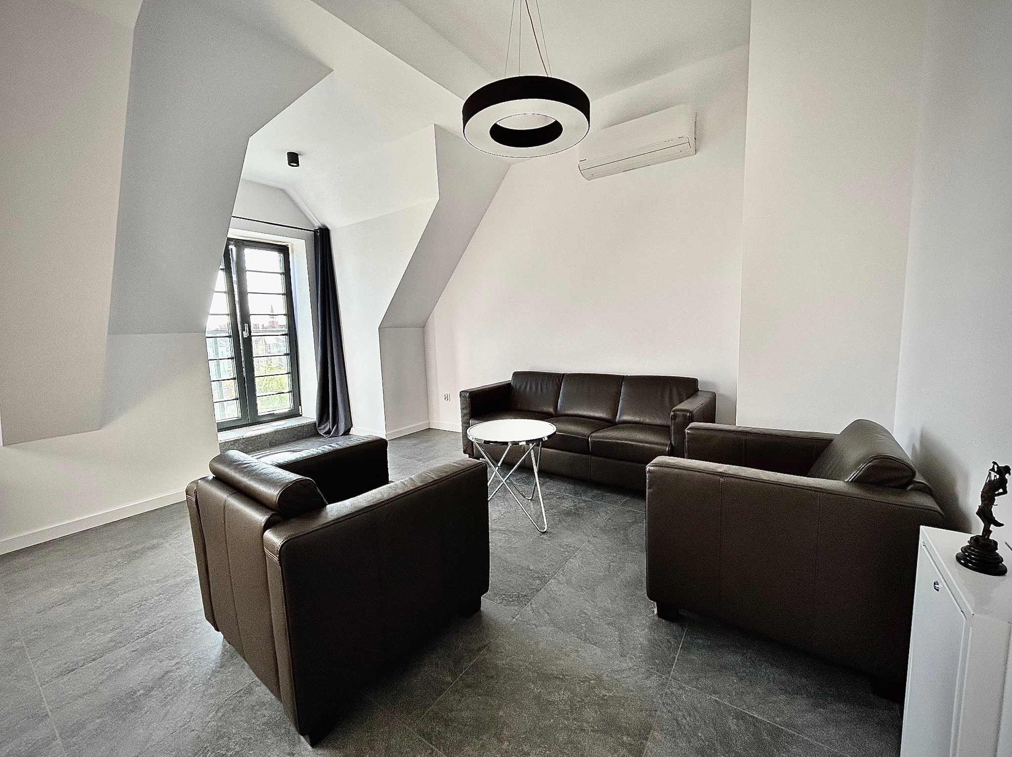 Klimatyzowane biuro kancelarii adwokackiej z brązowymi skórzanymi fotelami i sofą, okrągłym stolikiem na metalowych nogach, nowoczesną lampą sufitową i figurką Temidy na białej szafce.