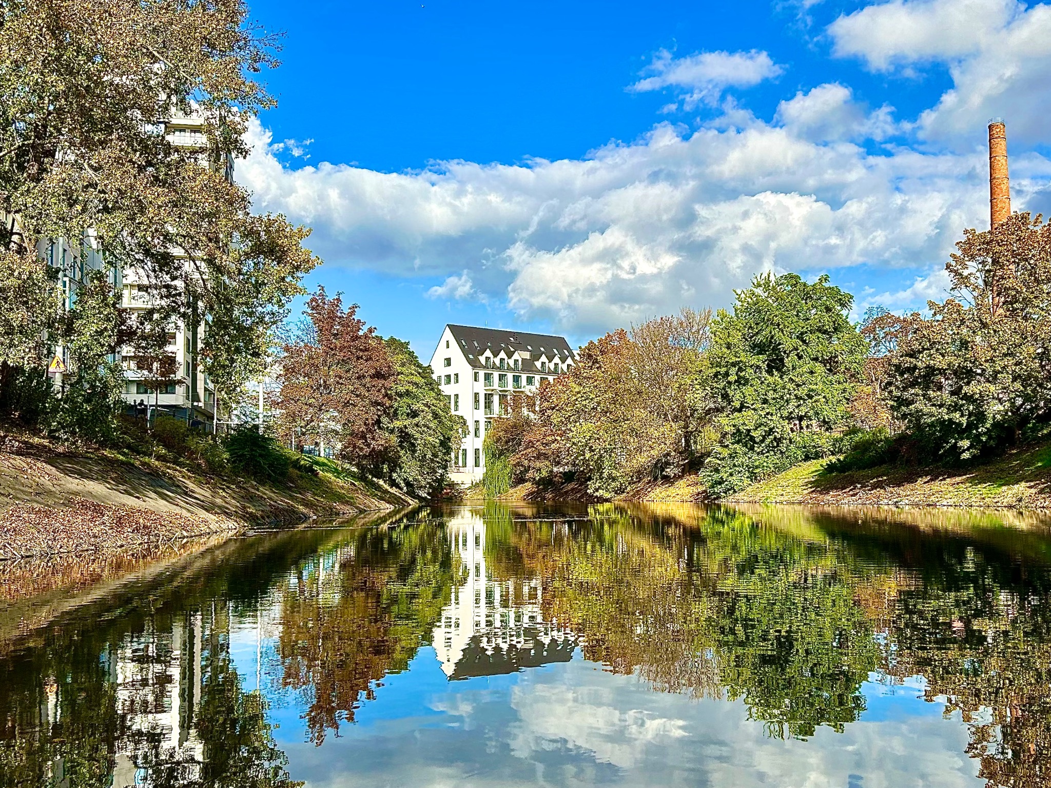Widok na biały budynek z czarnym dachem odbijający się w wodzie, otoczony drzewami w jesiennych kolorach i błękitnym niebem z chmurami.