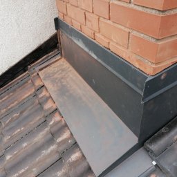 Usługi Blacharsko-Dekarskie oraz niewielkie prace ogolnobudowlane . Magdalena Zagaja - Szczegółowe ujęcie obróbki blacharskiej komina na dachu z brązowej dachówki ceramicznej, zakończonej szarą listwą, na tle ściany budynku.