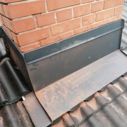 Usługi Blacharsko-Dekarskie oraz niewielkie prace ogolnobudowlane . Magdalena Zagaja - Szczegółowe ujęcie obróbki blacharskiej komina na dachu z dachówki ceramicznej. Ciemnoszara blacha, ceglany komin, widoczne zabrudzenia na dachówce.