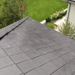 Usługi Blacharsko-Dekarskie oraz niewielkie prace ogolnobudowlane . Magdalena Zagaja - Nowy dach z ciemnej dachówki karpiówki, widok z góry na tle zieleni ogrodu i fragmentu budynku. Dach świeżo położony, czysty, bez uszkodzeń.