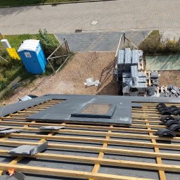 Usługi Blacharsko-Dekarskie oraz niewielkie prace ogolnobudowlane . Magdalena Zagaja - Montaż dachówki ceramicznej na drewnianej konstrukcji dachu. Widoczny fragment okna dachowego i zapas dachówek. Na drugim planie toaleta przenośna i palety z materiałem.