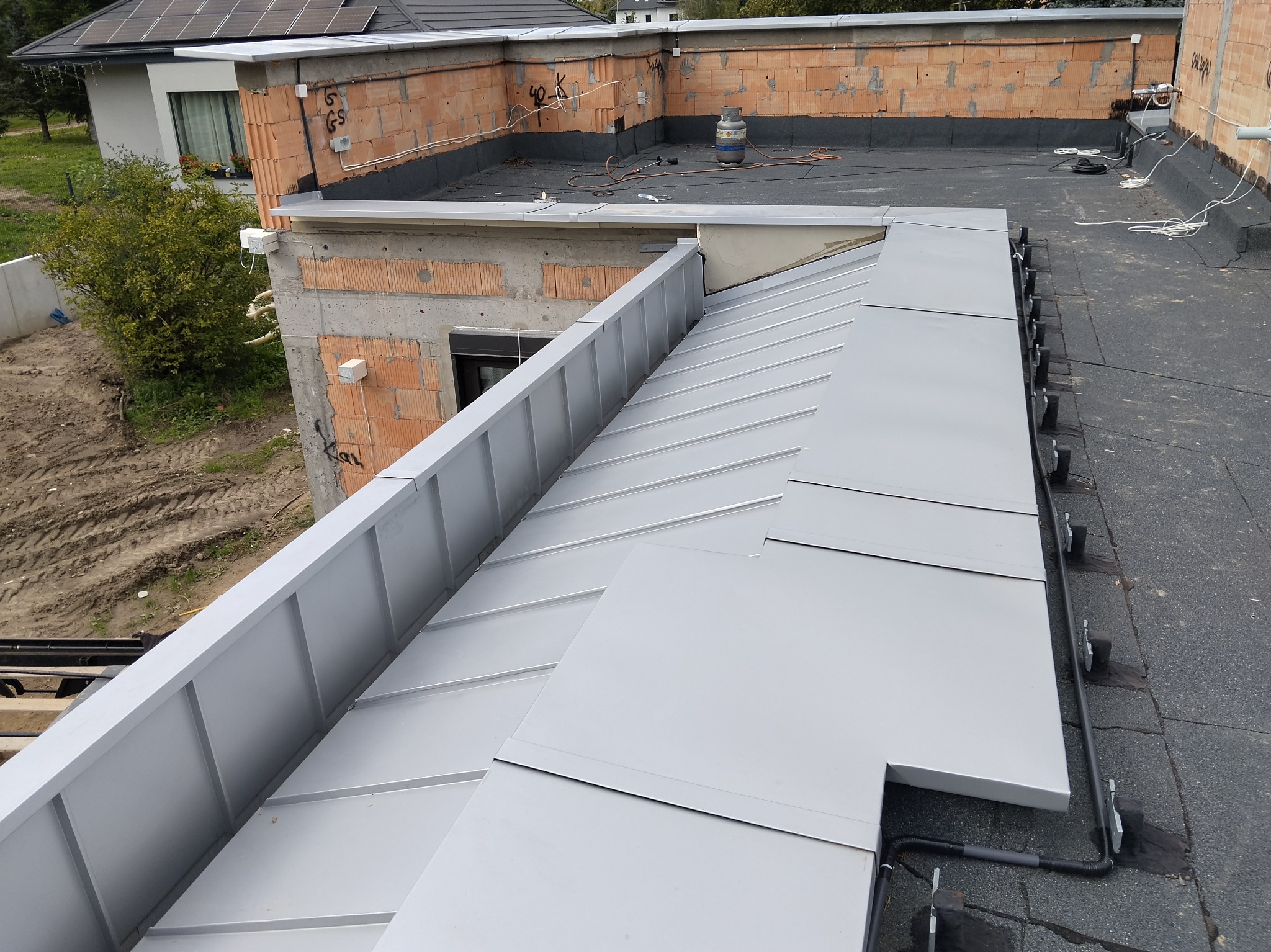 Nowoczesny dach płaski z blachy trapezowej w odcieniach szarości, widok z góry, z elementami instalacji i niezakończonymi ścianami budynku w tle.