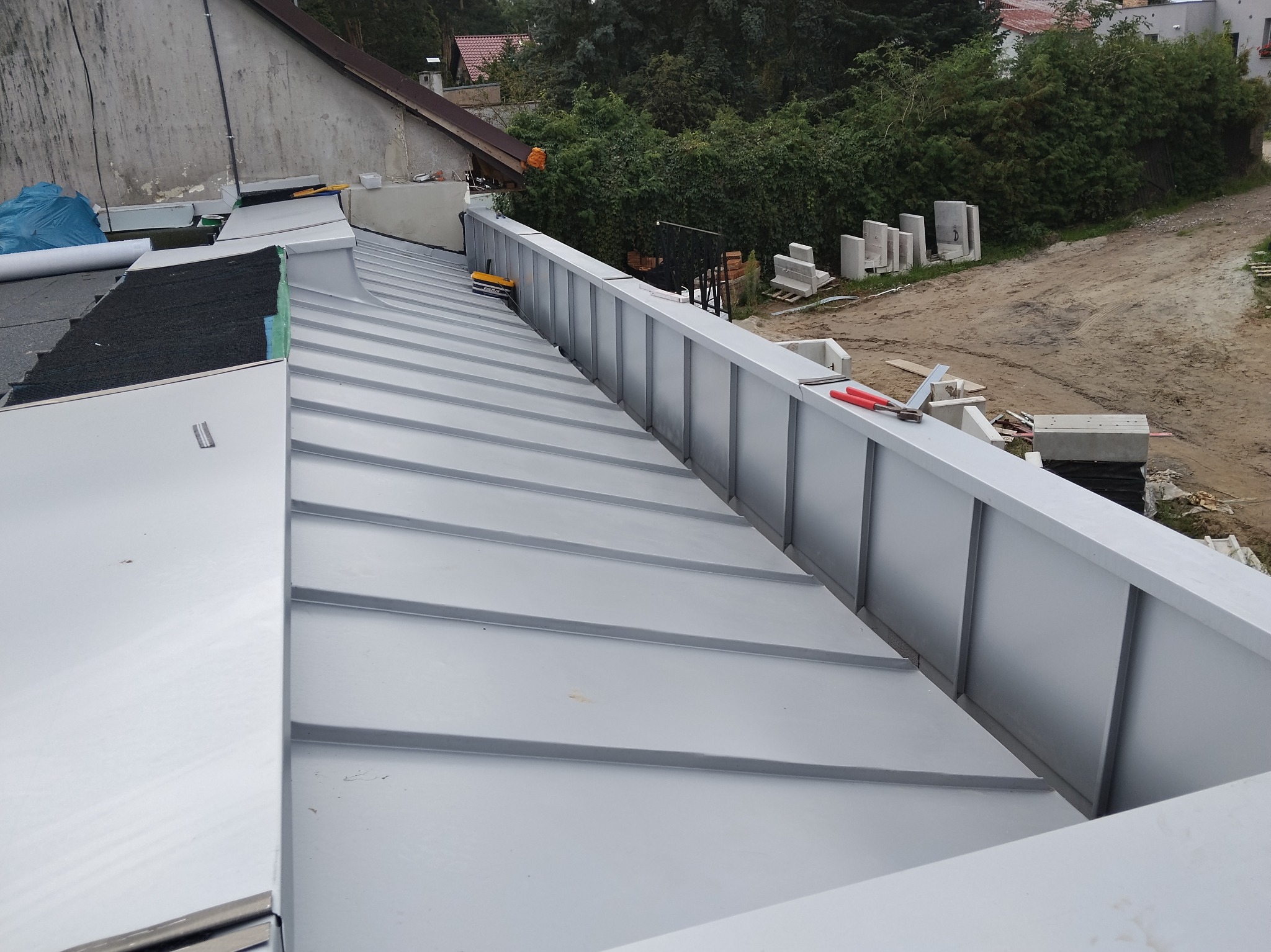 Szary dach z blachy trapezowej w trakcie montażu, widoczne narzędzia i elementy konstrukcyjne. Tło stanowią budynki i drzewa. Ujęcie z góry.