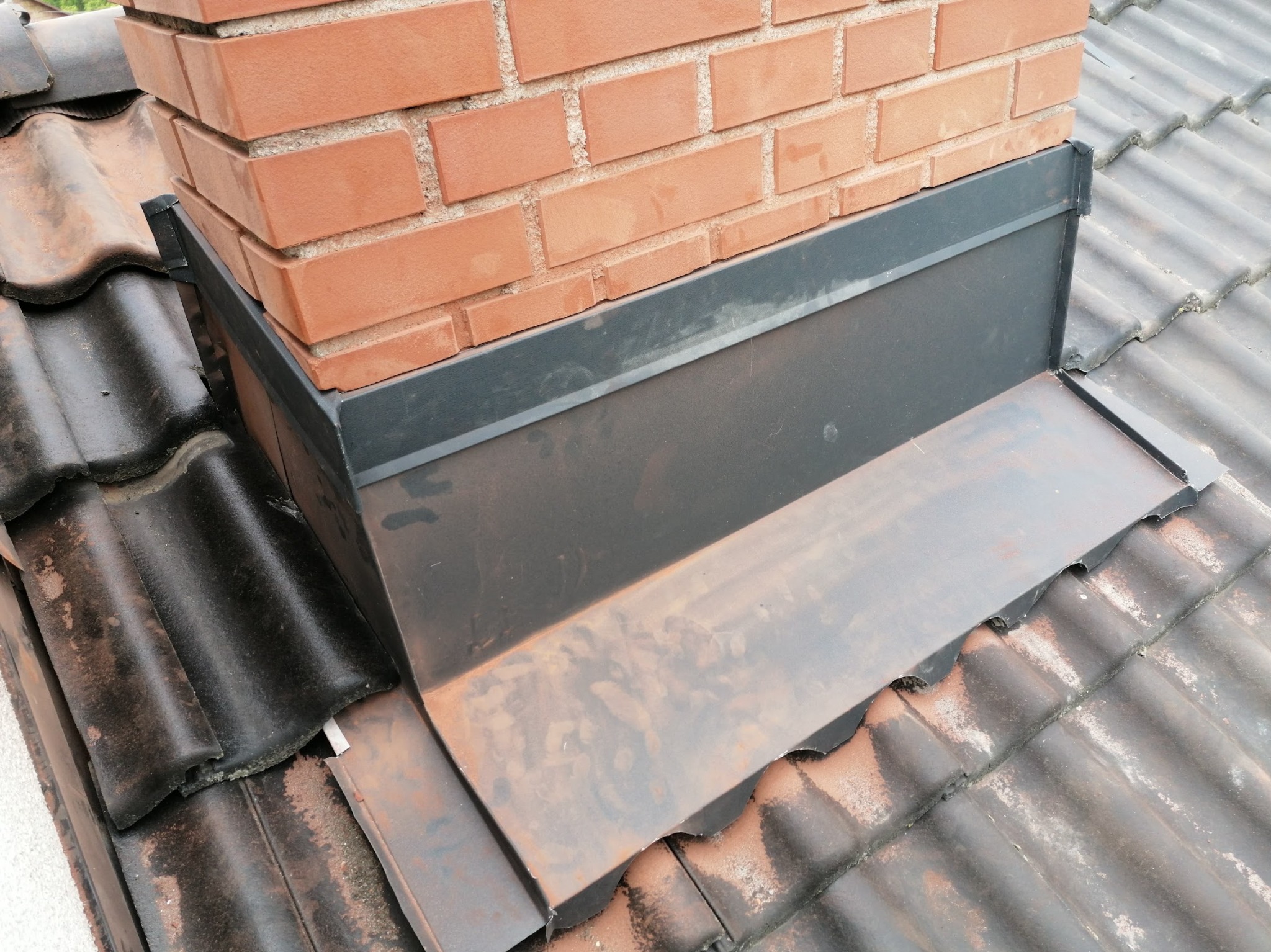 Szczegółowe ujęcie obróbki blacharskiej komina na dachu z dachówki ceramicznej. Ciemnoszara blacha, ceglany komin, widoczne zabrudzenia na dachówce.