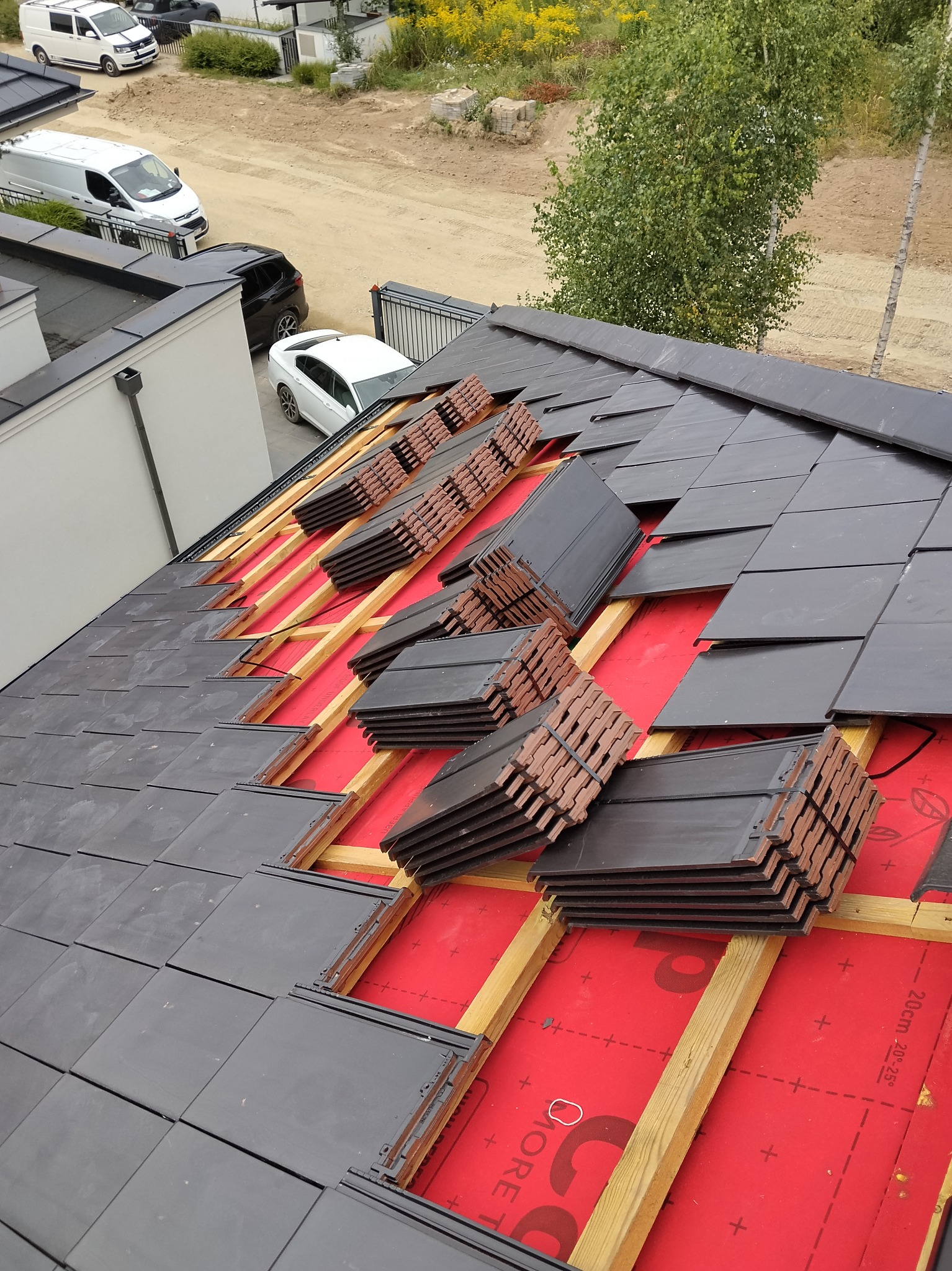 Montaż dachówki ceramicznej na czerwonej membranie dachowej. Częściowo pokryty dach z widocznymi stertami dachówek do ułożenia. Widok z góry, w tle zabudowa jednorodzinna.
