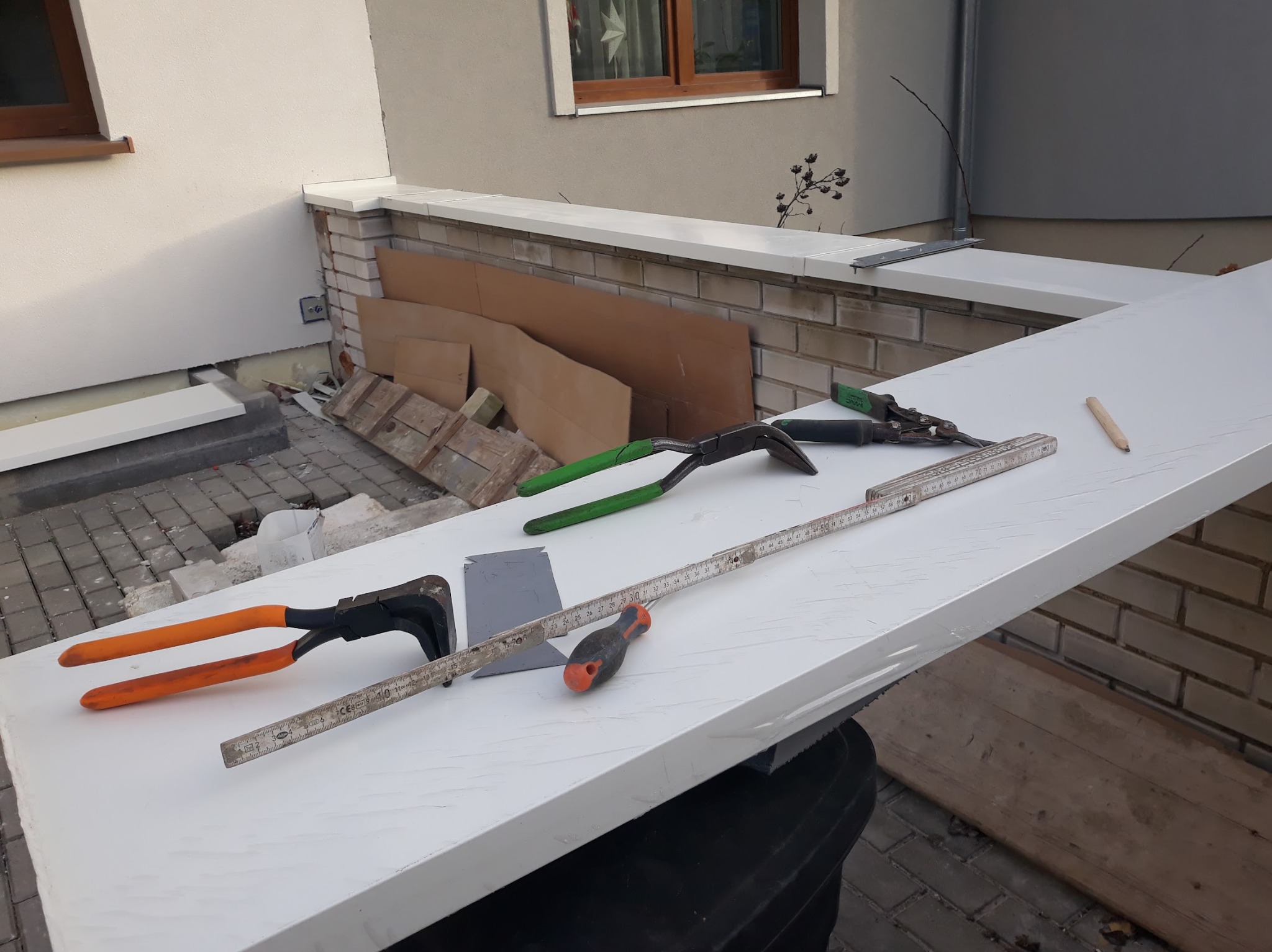 Biały element wykończeniowy z narzędziami blacharskimi na tle budynku. Widoczne cęgi, miarka i ołówek.