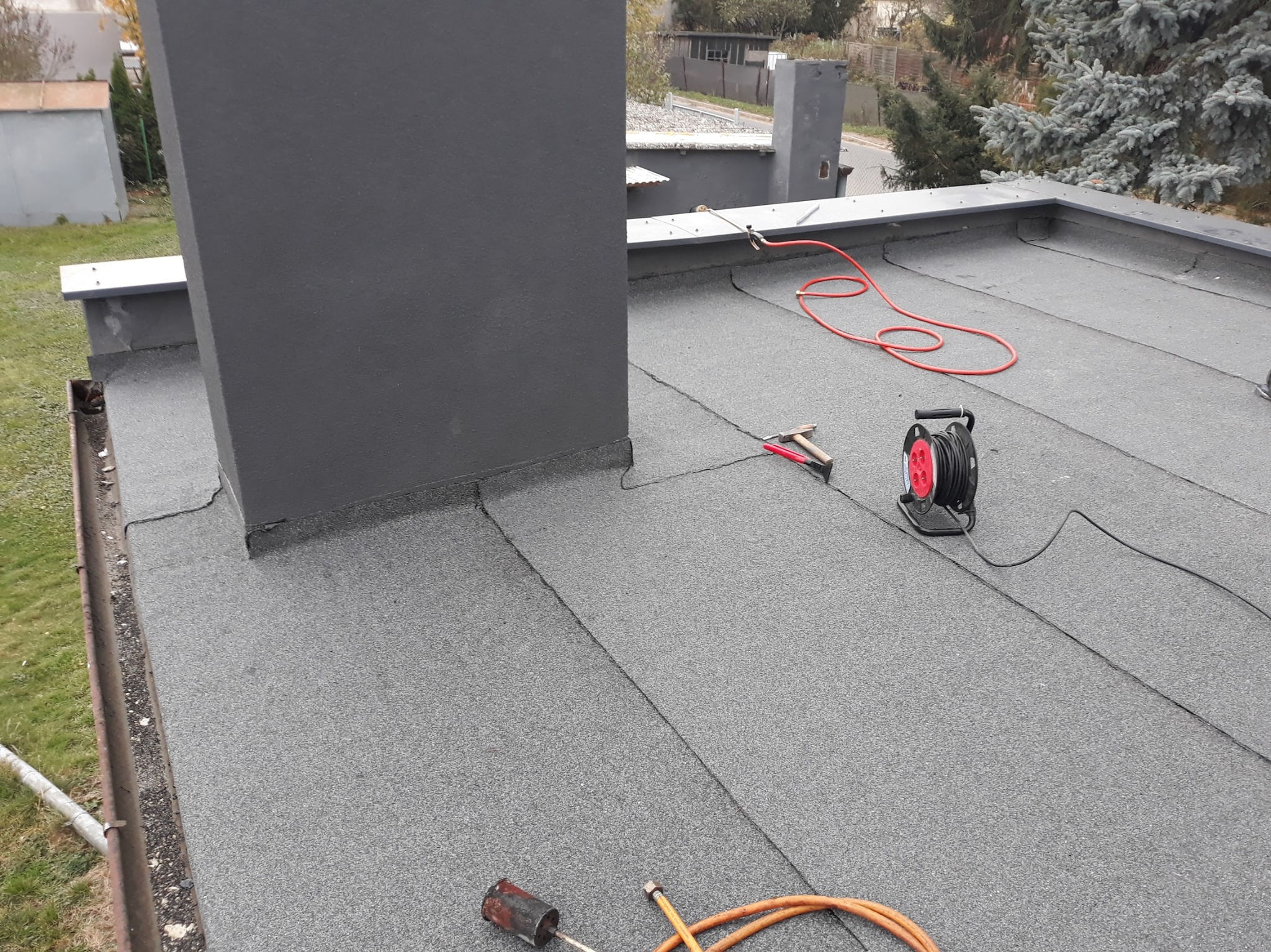 Szary, płaski dach pokryty papą, z kominem i narzędziami dekarskimi. Widoczna nowa izolacja wokół komina i narzędzia do zgrzewania papy.