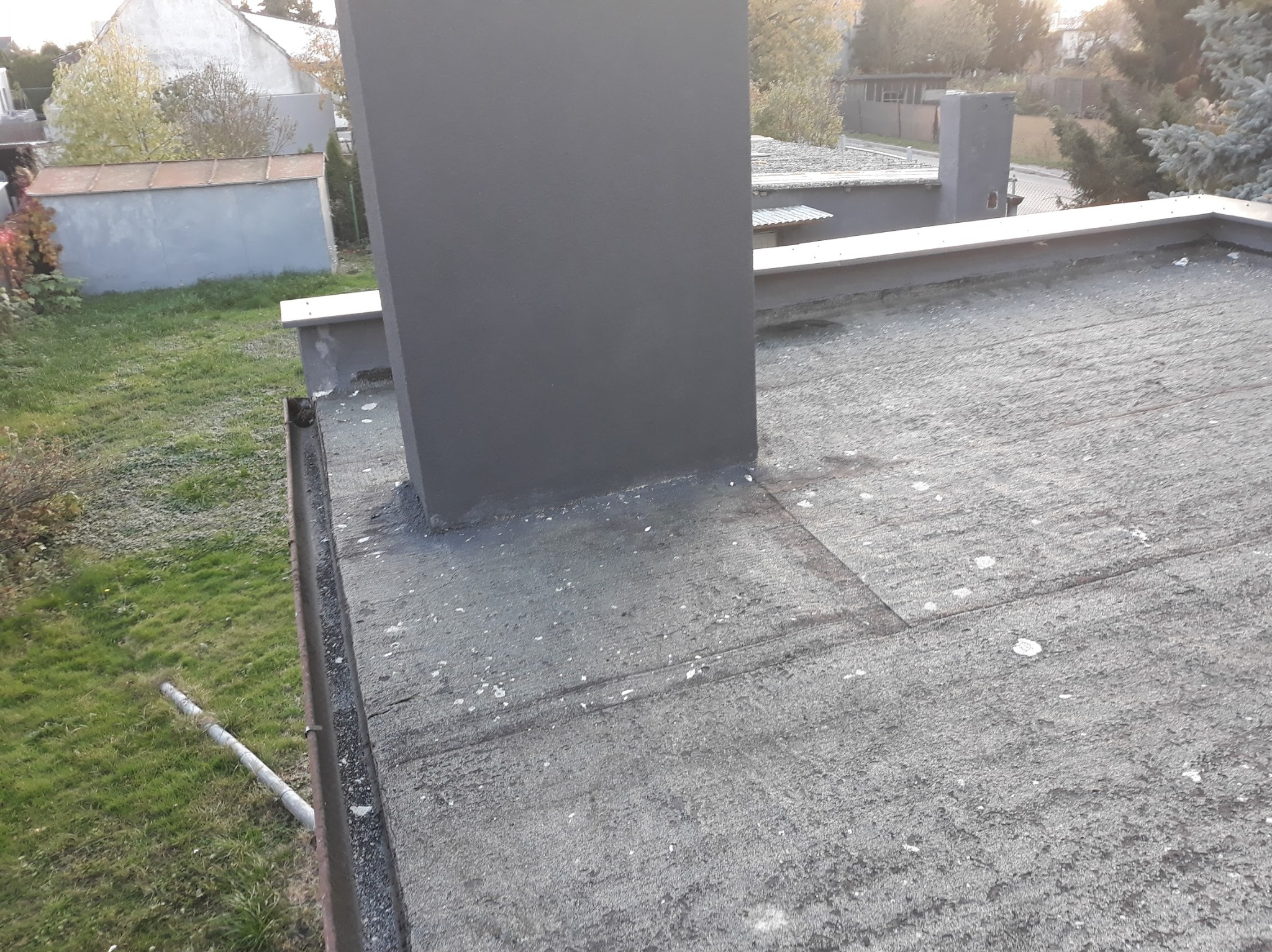 Szczegółowe ujęcie płaskiego dachu z papy, z kominem w kolorze grafitowym. Widoczne obróbki blacharskie i fragment otoczenia z zielenią. Dach pokryty białymi plamkami.