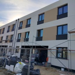 VOLODYMYR IVANOV - Trzykondygnacyjny budynek w trakcie prac elewacyjnych, widoczne rusztowania, ocieplenie części ściany w odcieniach beżu i bieli, czarne ramy okienne, teren budowy z workami i materiałami.