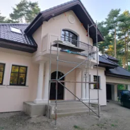 Elewacja domu w odcieniach beżu z ciemną stolarką okienną, widoczne rusztowanie, brązowy dach i otoczenie drzew. Prace wykończeniowe.