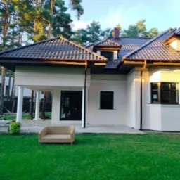 Elewacja domu jednorodzinnego w jasnym kolorze, z brązowym dachem i elementami wykończeniowymi. W tle drzewa iglaste i trawnik z rattanowym fotelem.