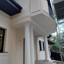 Jasna elewacja domu z balkonem, ozdobiona kolumnami i sztukaterią. Widoczne elementy wykończeniowe i rusztowanie. Detal architektoniczny z bliska.