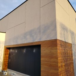 VOLODYMYR IVANOV - Nowoczesna elewacja domu z jasnoszarym tynkiem i drewnianą okładziną nad garażem. Widoczne detale łączeń płyt elewacyjnych i cień drzewa na ścianie.