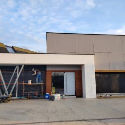 VOLODYMYR IVANOV - Pracownik na rusztowaniu maluje fragment elewacji nowoczesnego domu z elementami drewna i szarego tynku. Widoczne narzędzia i materiały budowlane.