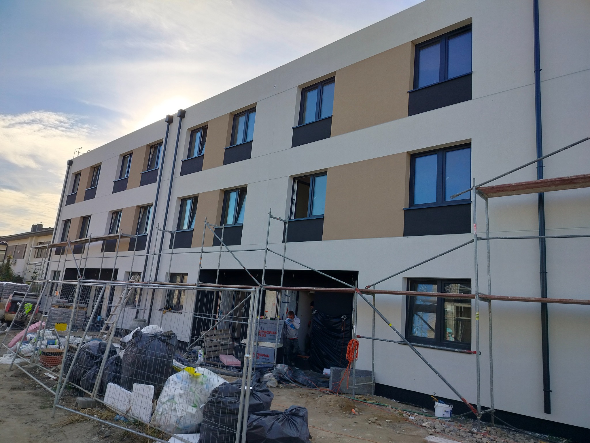 Trzykondygnacyjny budynek w trakcie prac elewacyjnych, widoczne rusztowania, ocieplenie części ściany w odcieniach beżu i bieli, czarne ramy okienne, teren budowy z workami i materiałami.