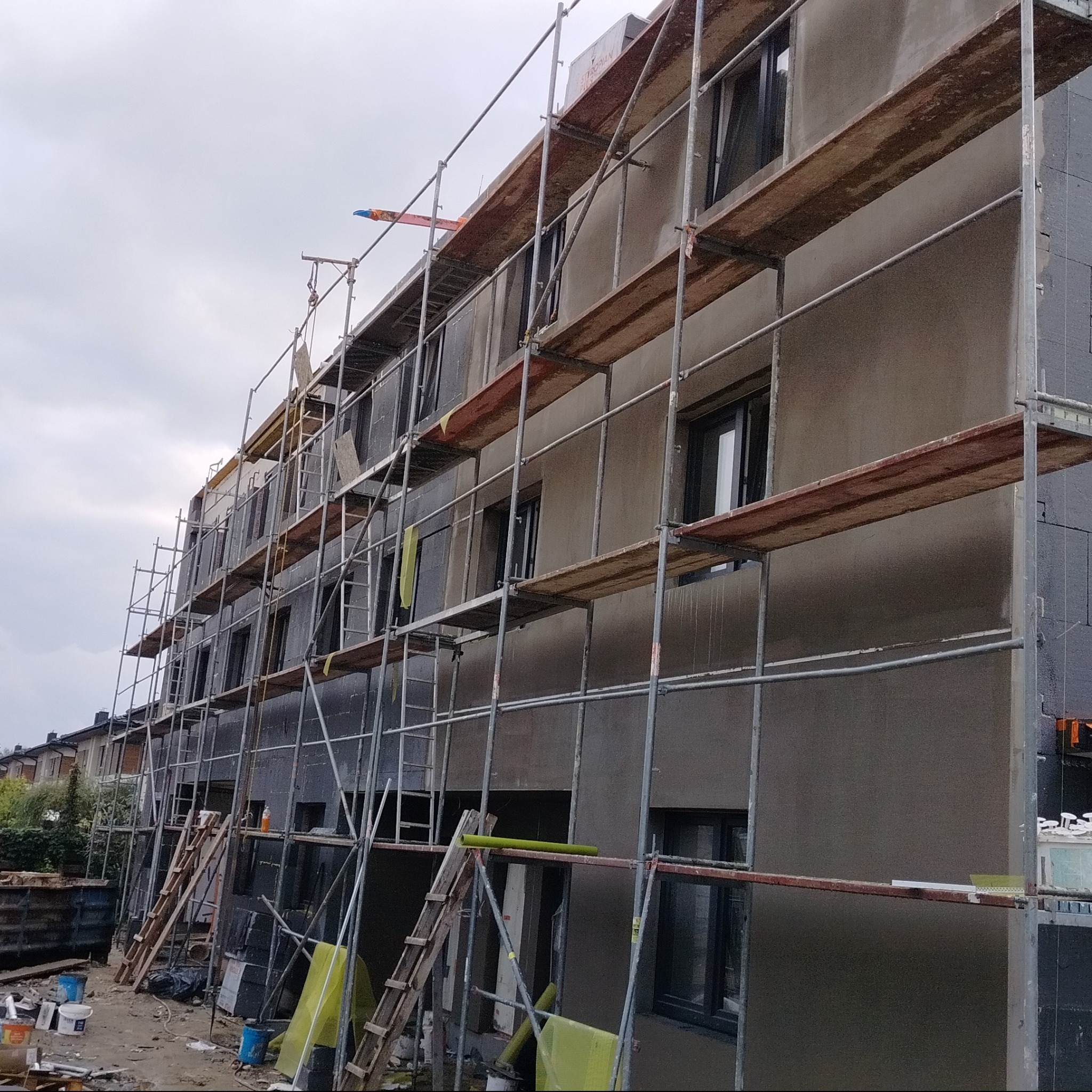 Wielopiętrowy budynek w trakcie tynkowania elewacji, widoczne rusztowania, częściowo otynkowana szara ściana, okna z czarnymi ramami, drabiny i materiały budowlane na dole.