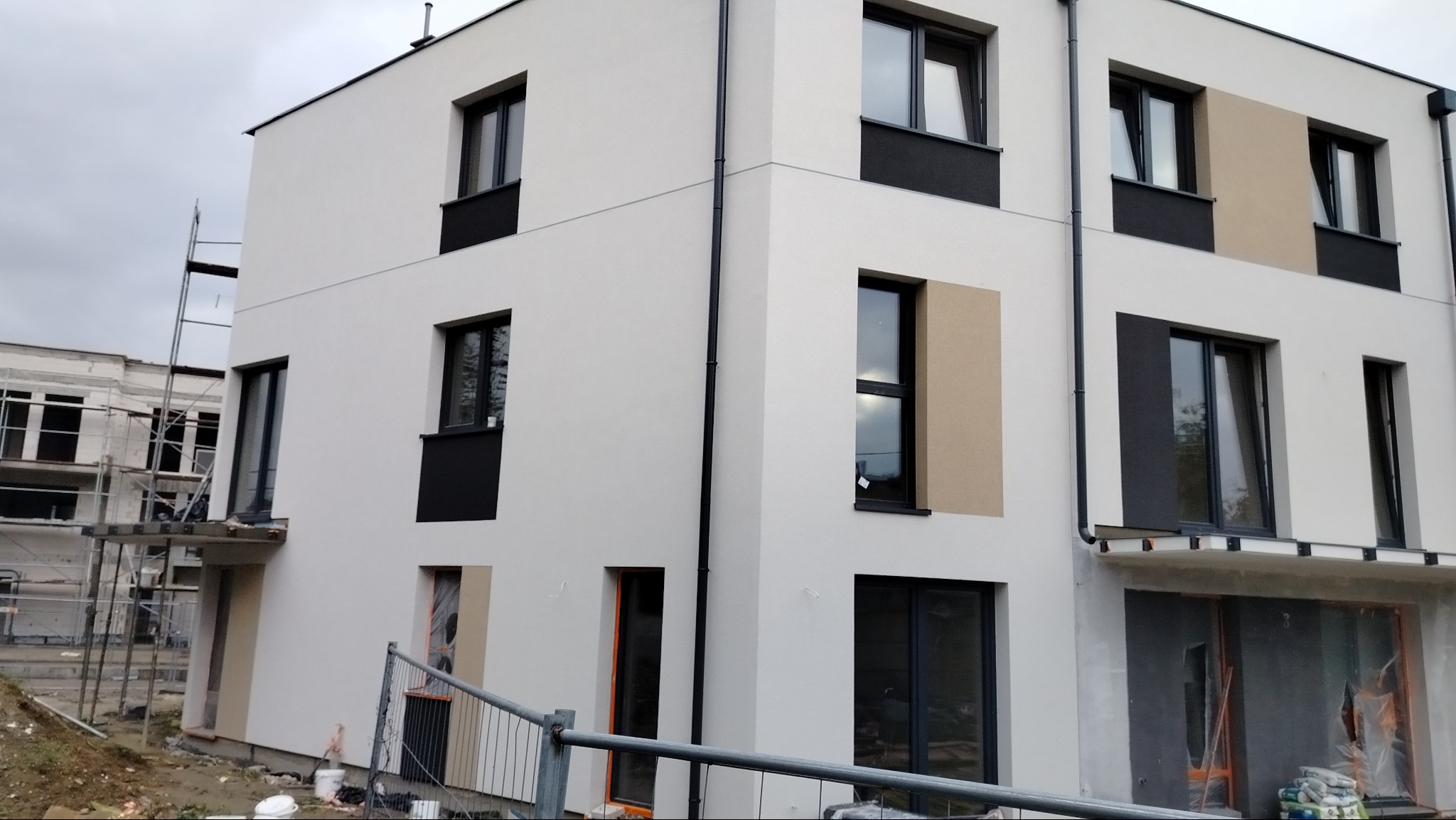 Nowoczesny, trzypiętrowy budynek z białą elewacją, czarnymi ramami okiennymi i akcentami w kolorze piaskowym, widoczny fragment rusztowania i teren budowy.