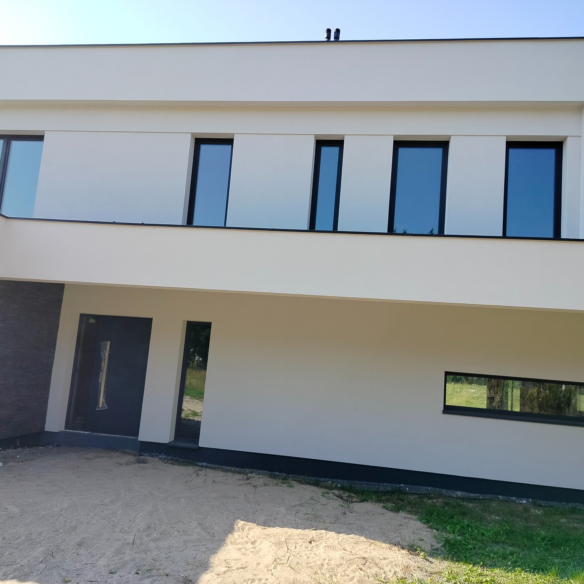 Nowoczesny dom z białą elewacją, czarnymi ramami okiennymi i drzwiami, widoczne dwa kominy na dachu, teren wokół domu częściowo pokryty piaskiem.