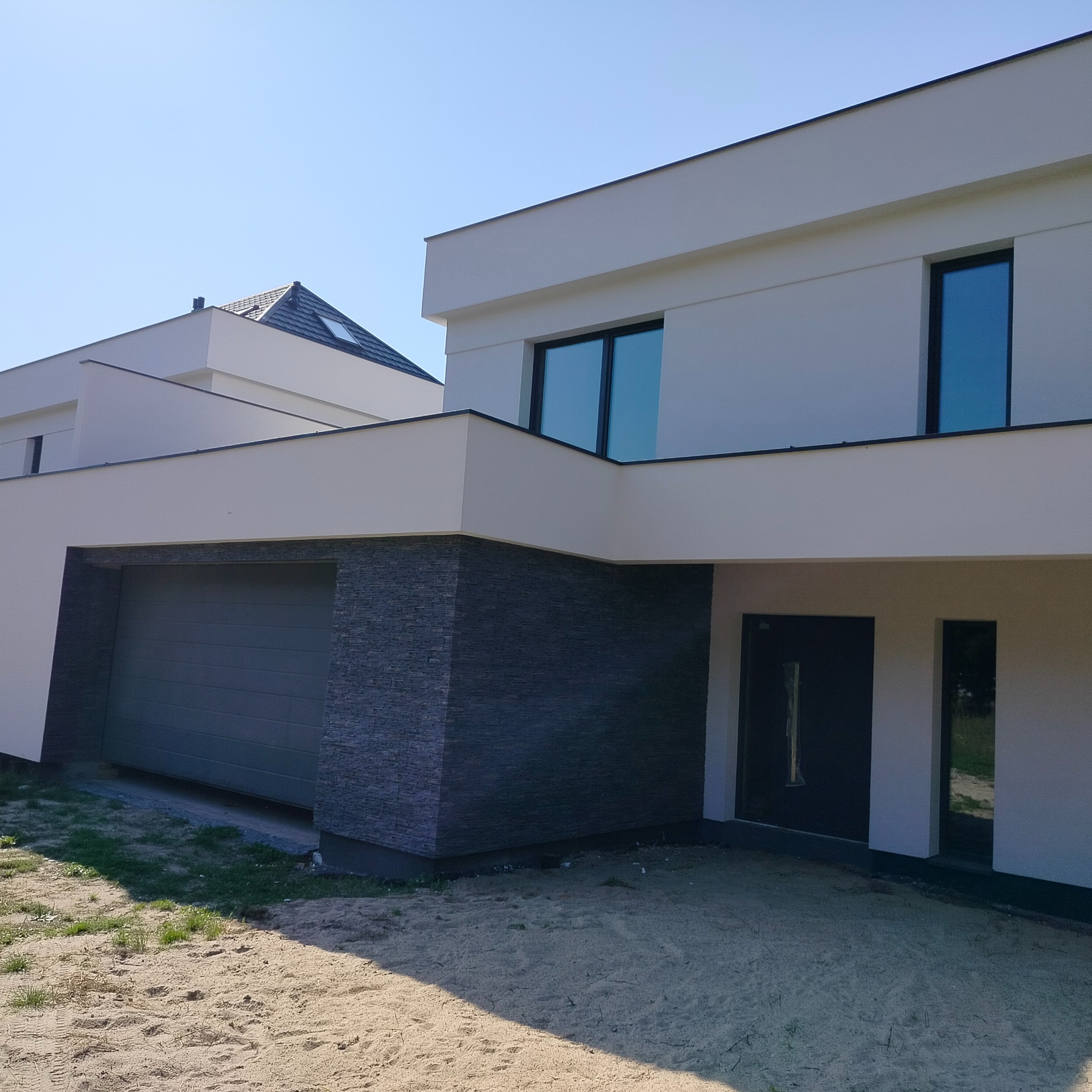 Nowoczesny dom jednorodzinny z jasną elewacją, grafitową okładziną przy garażu i ciemną stolarką okienną, widok z zewnątrz na tle błękitnego nieba.