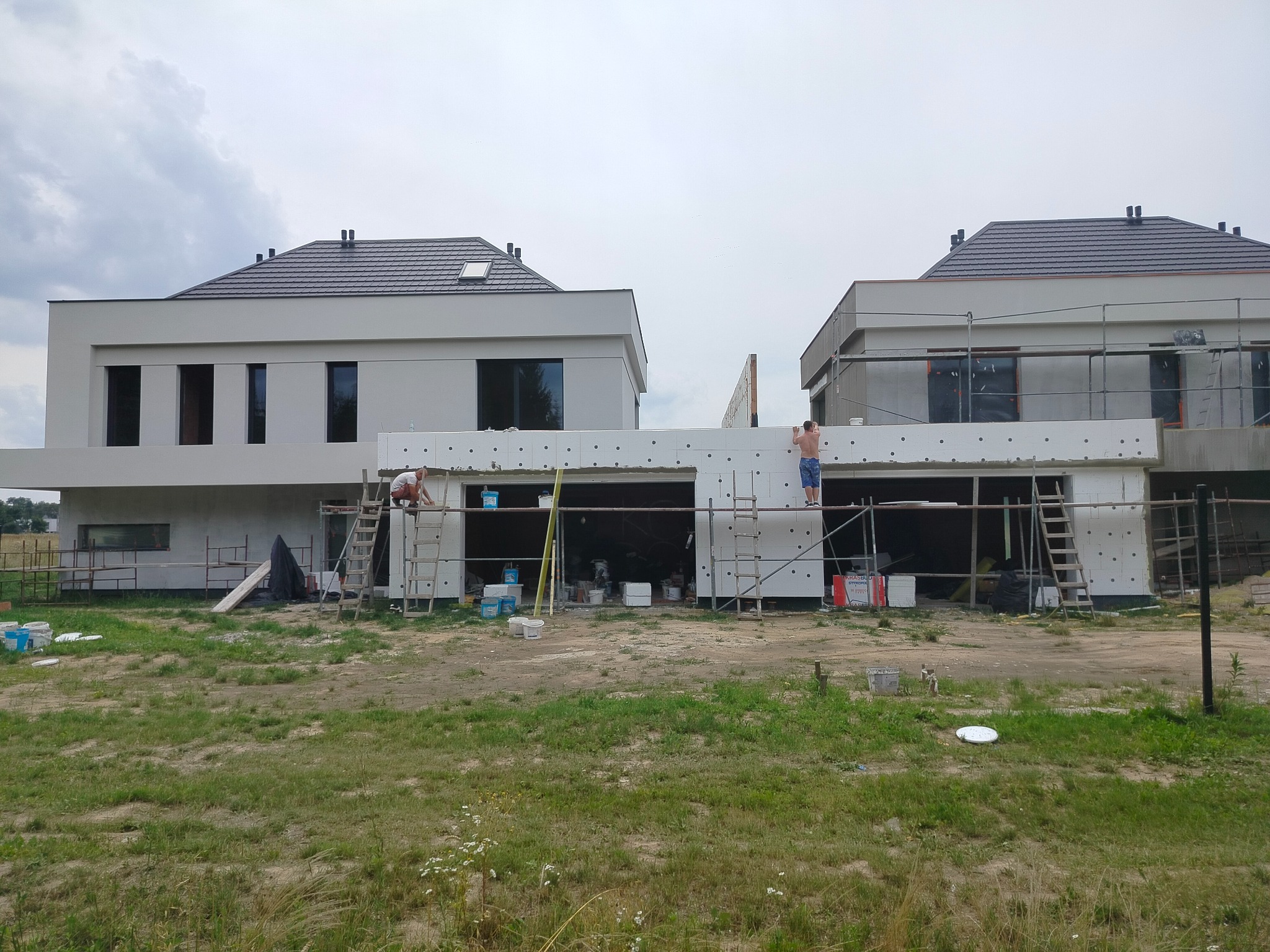 Dwa nowoczesne, jasne domy w trakcie ocieplania styropianem, widoczne rusztowania i pracownicy w trakcie montażu płyt izolacyjnych, teren wokół w stanie surowym.