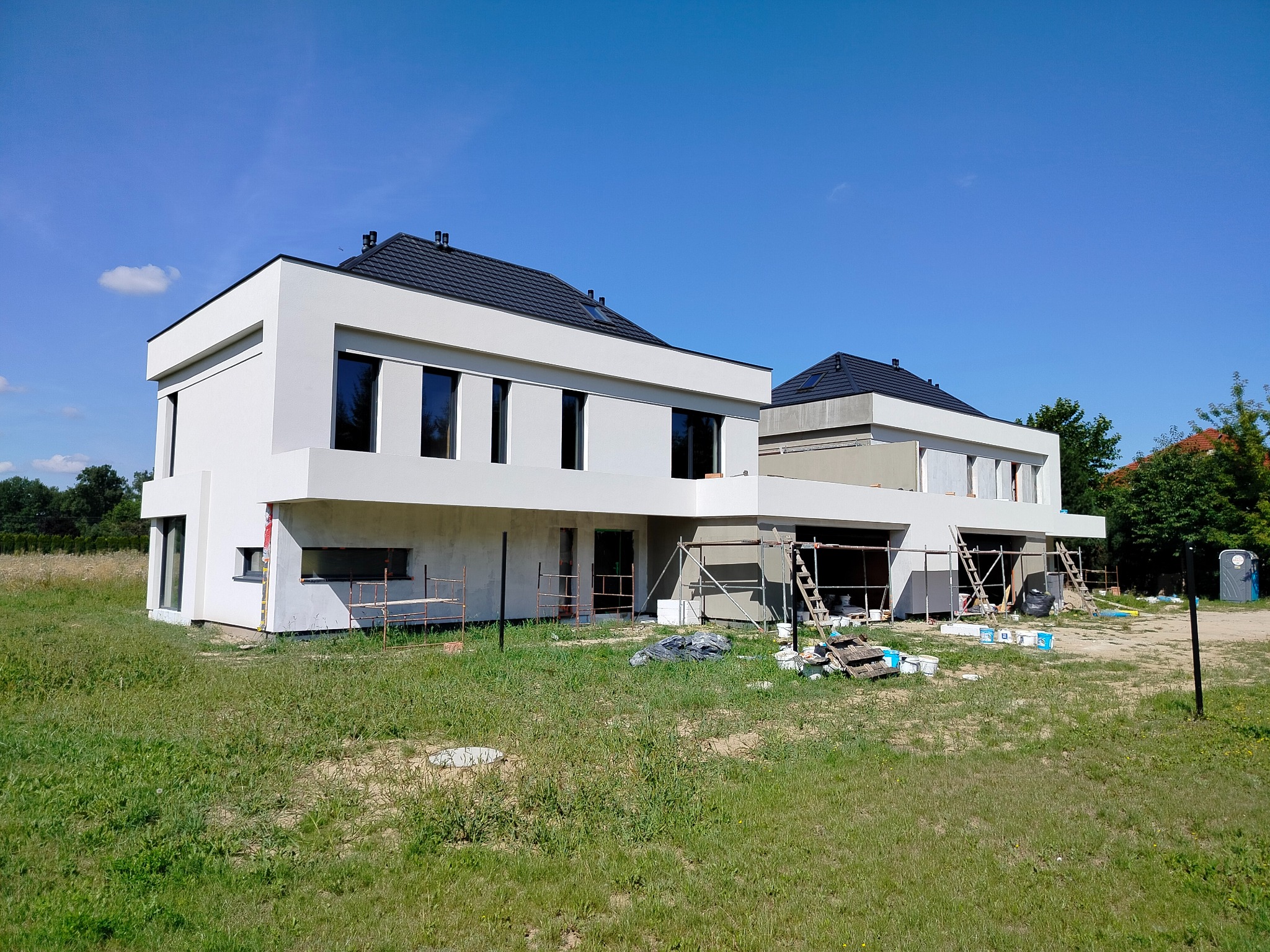Dwa nowoczesne domy w trakcie wykańczania elewacji zewnętrznej, widoczne rusztowania, puszki z farbą i materiały budowlane na trawniku.