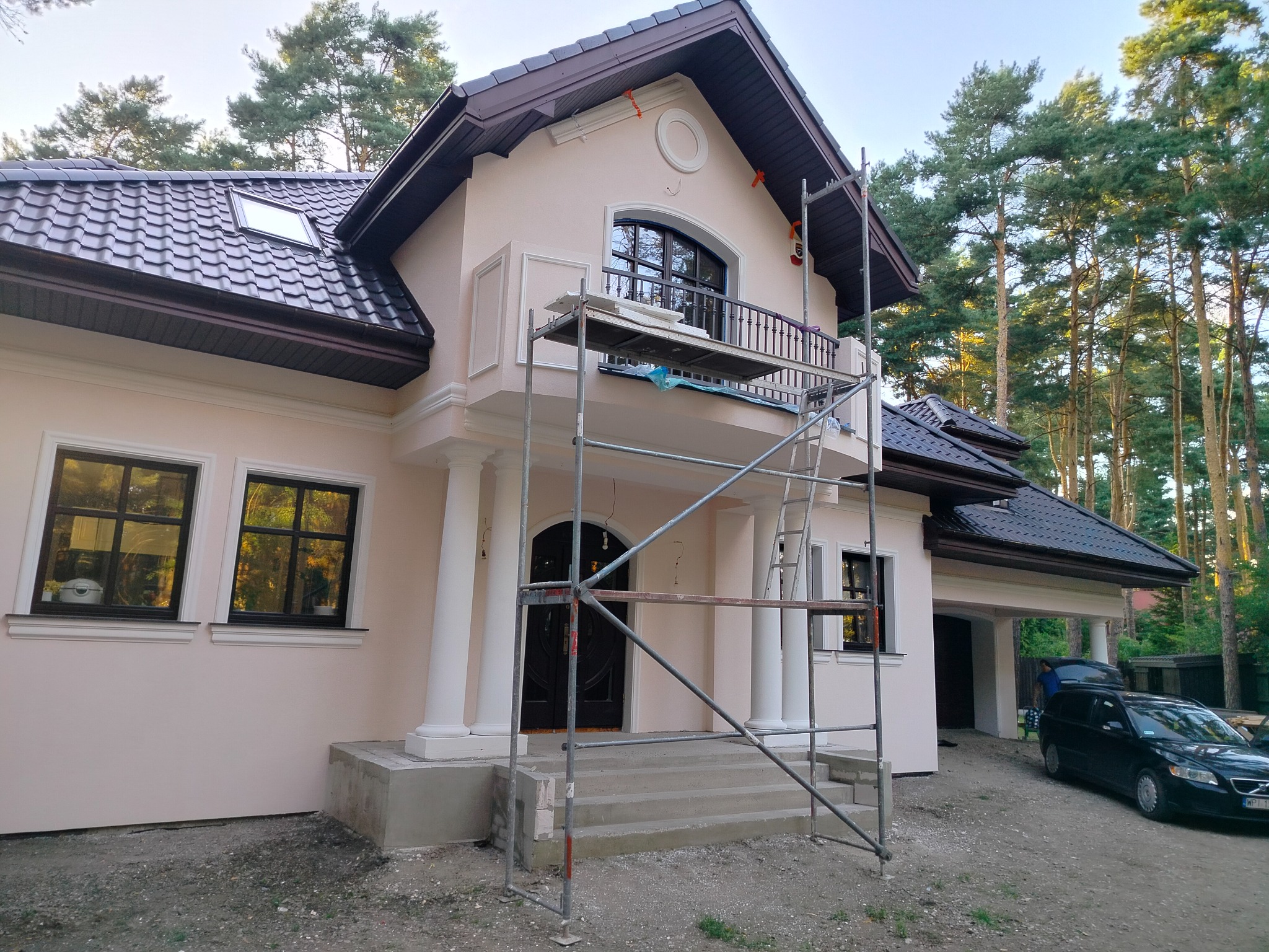 Elewacja domu w odcieniach beżu z ciemną stolarką okienną, widoczne rusztowanie, brązowy dach i otoczenie drzew. Prace wykończeniowe.