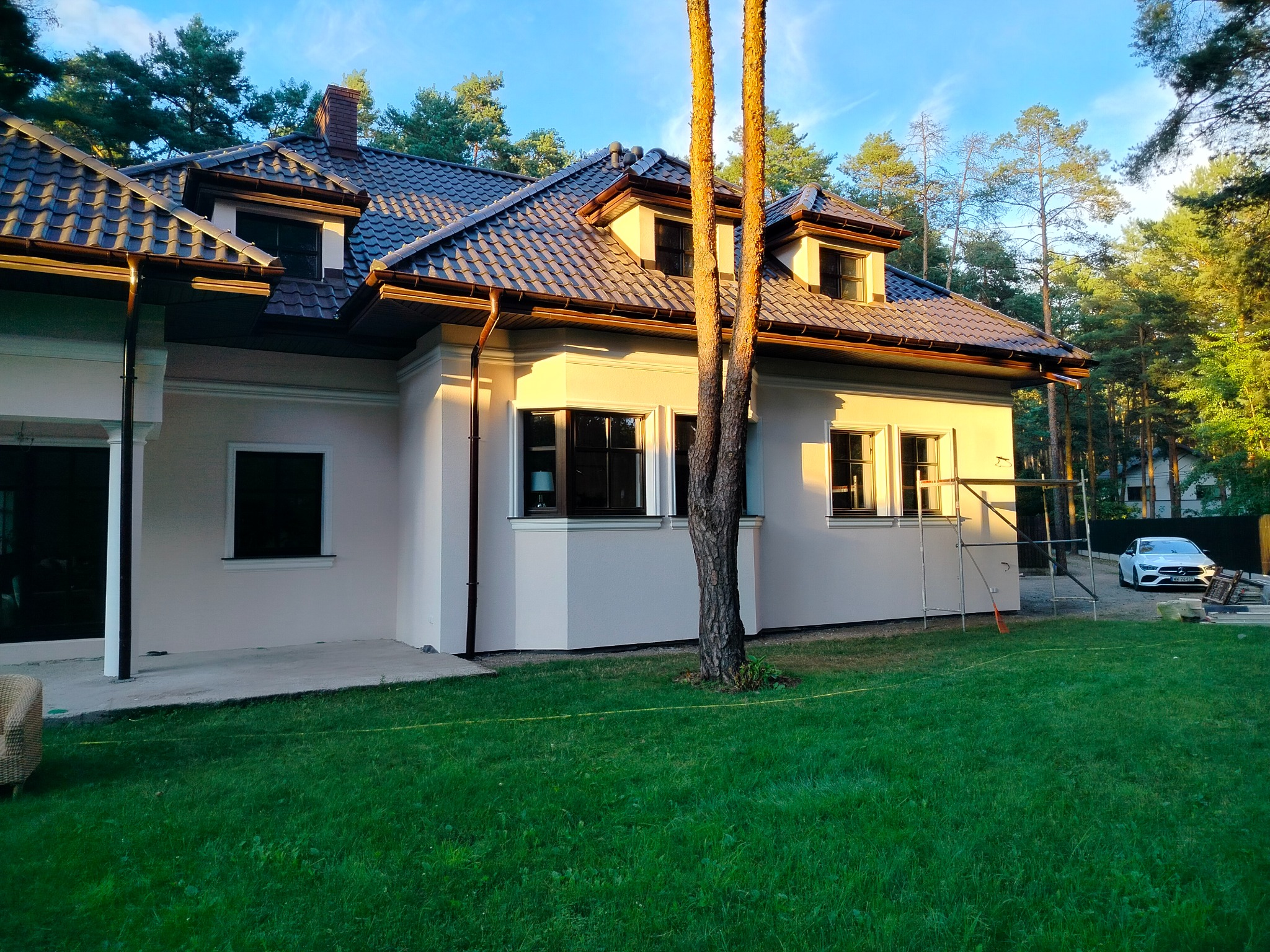 Jasna elewacja domu jednorodzinnego z ciemną dachówką, widoczna zieleń trawnika i drzewa w tle, rusztowanie obok ściany, ciepłe światło słoneczne.