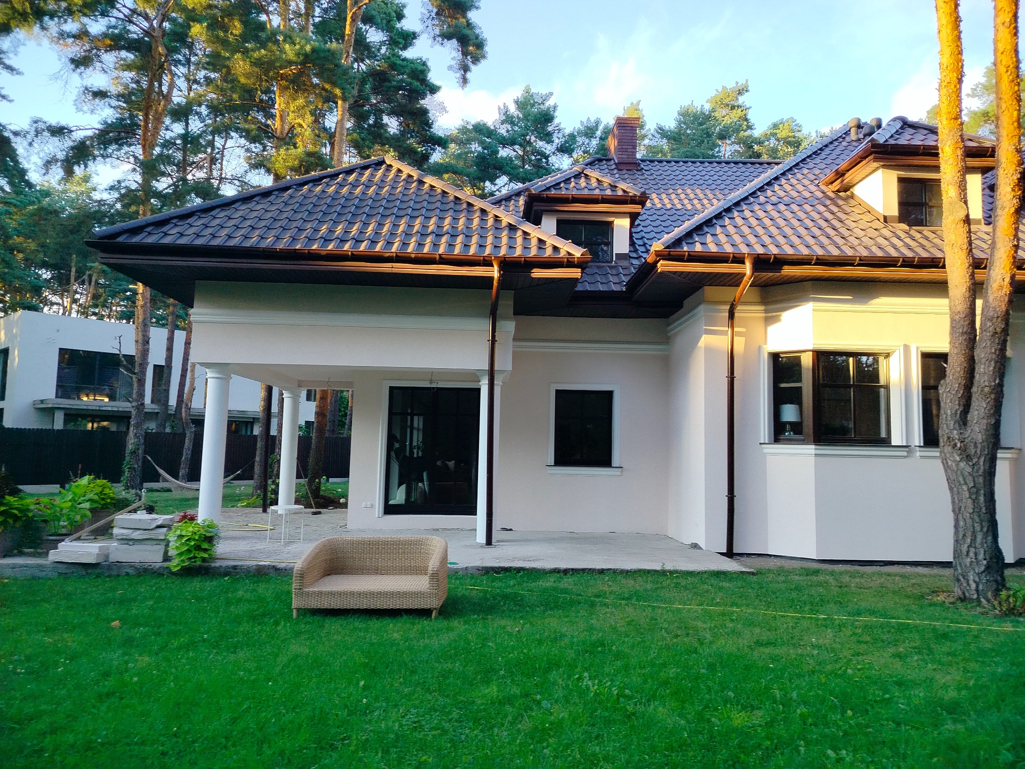 Elewacja domu jednorodzinnego w jasnym kolorze, z brązowym dachem i elementami wykończeniowymi. W tle drzewa iglaste i trawnik z rattanowym fotelem.