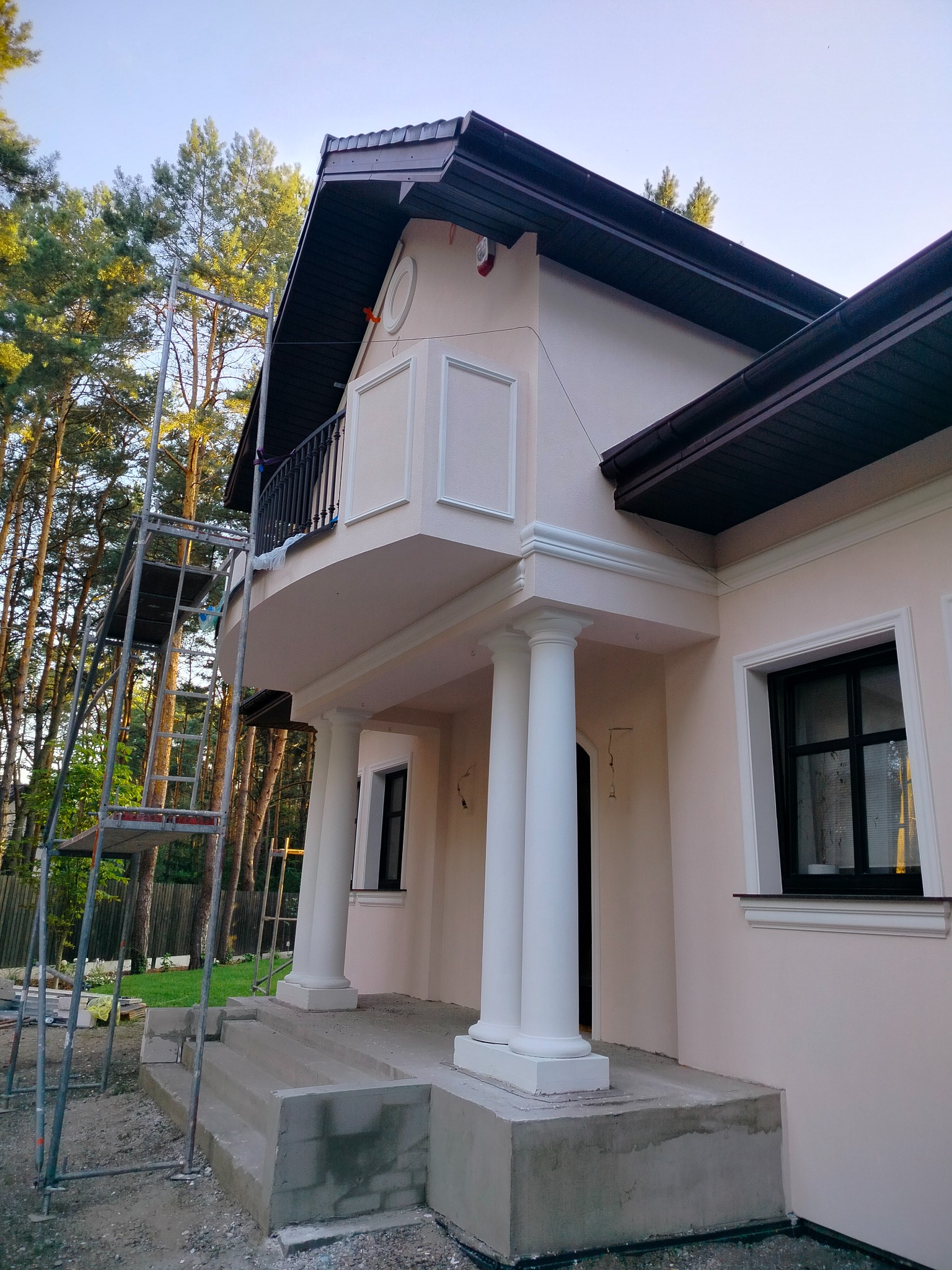 Jasna elewacja domu z kolumnami i balkonem, widoczne rusztowania. Detale architektoniczne podkreślone bielą, otoczenie drzewa. Ukończony etap prac.