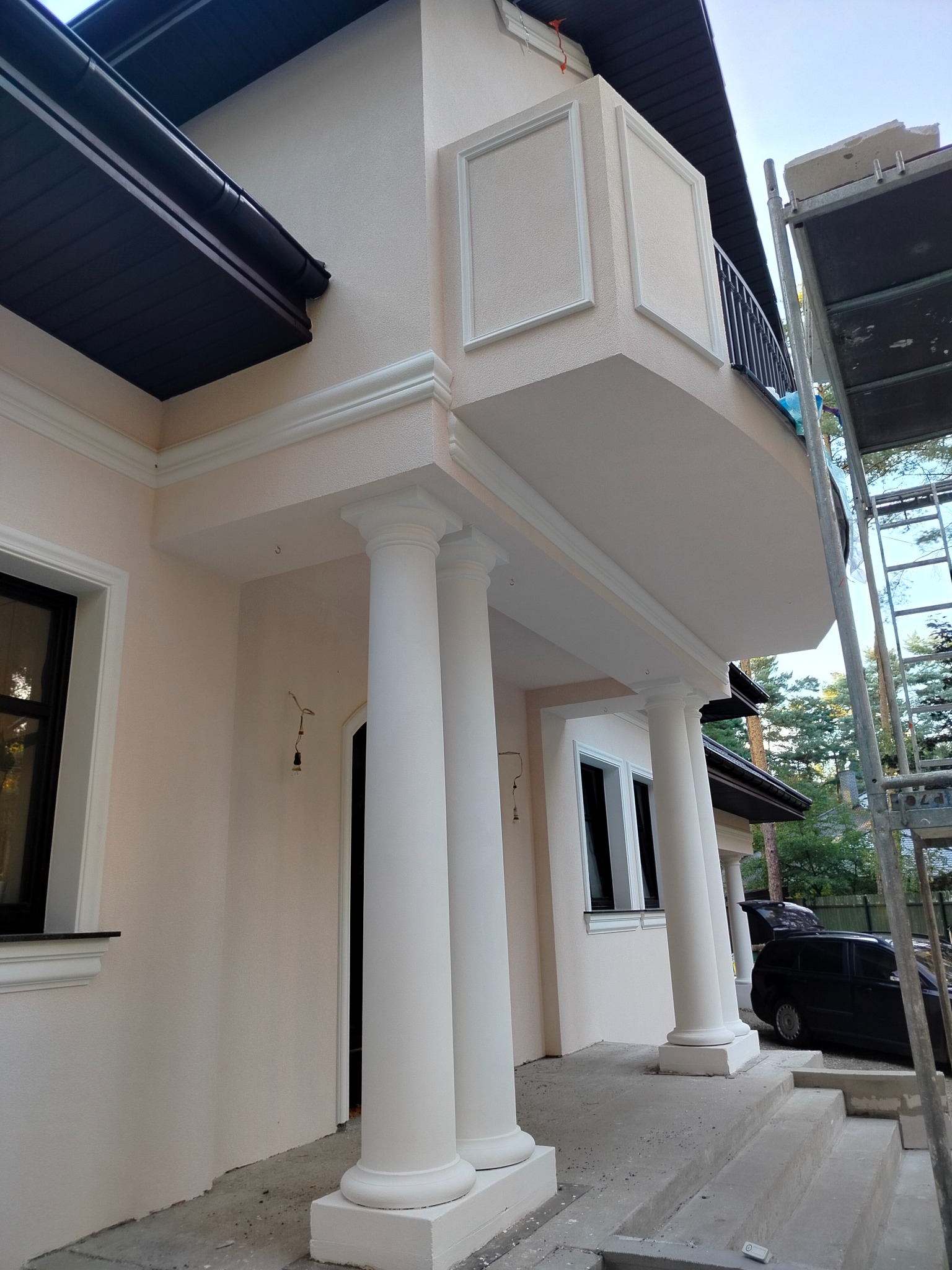 Jasna elewacja domu z balkonem, ozdobiona kolumnami i sztukaterią. Widoczne elementy wykończeniowe i rusztowanie. Detal architektoniczny z bliska.