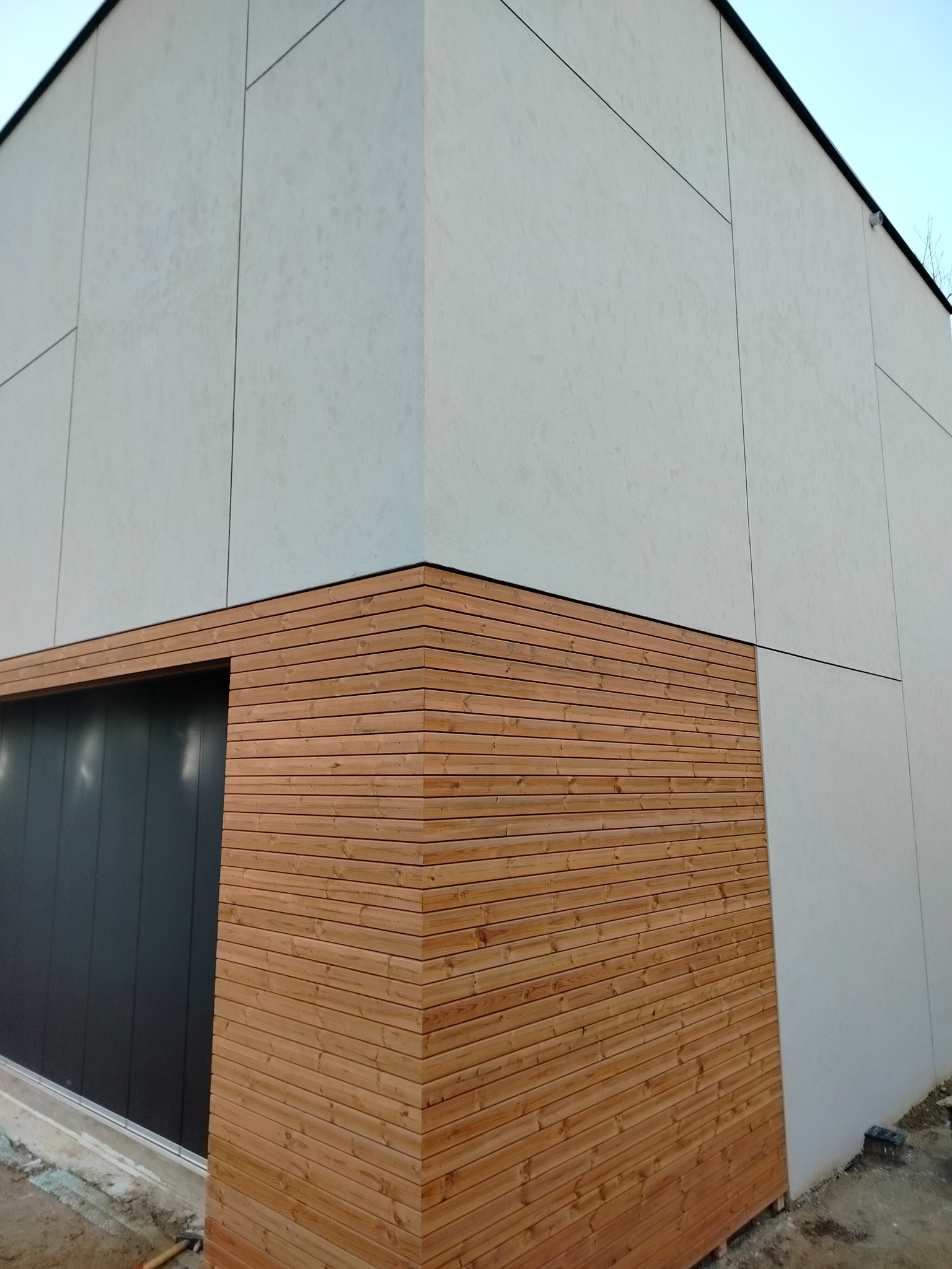 Nowoczesna elewacja budynku z jasnoszarymi panelami i drewnianą okładziną. Widoczny fragment bramy garażowej. Minimalistyczny design.