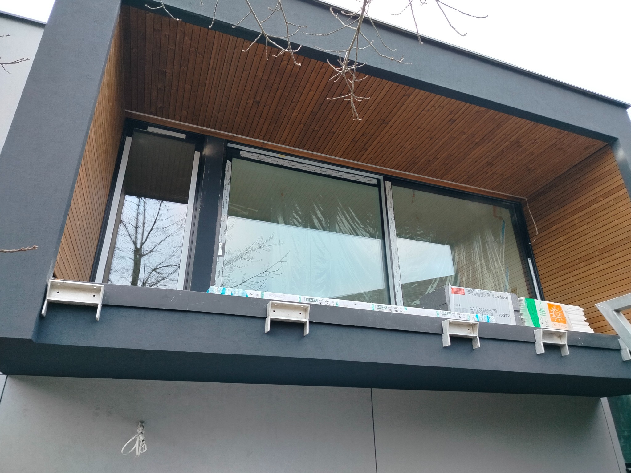 Nowoczesna elewacja domu z drewnianym podbitką i dużym oknem, widoczne elementy montażowe i materiały izolacyjne, szara kolorystyka ścian.