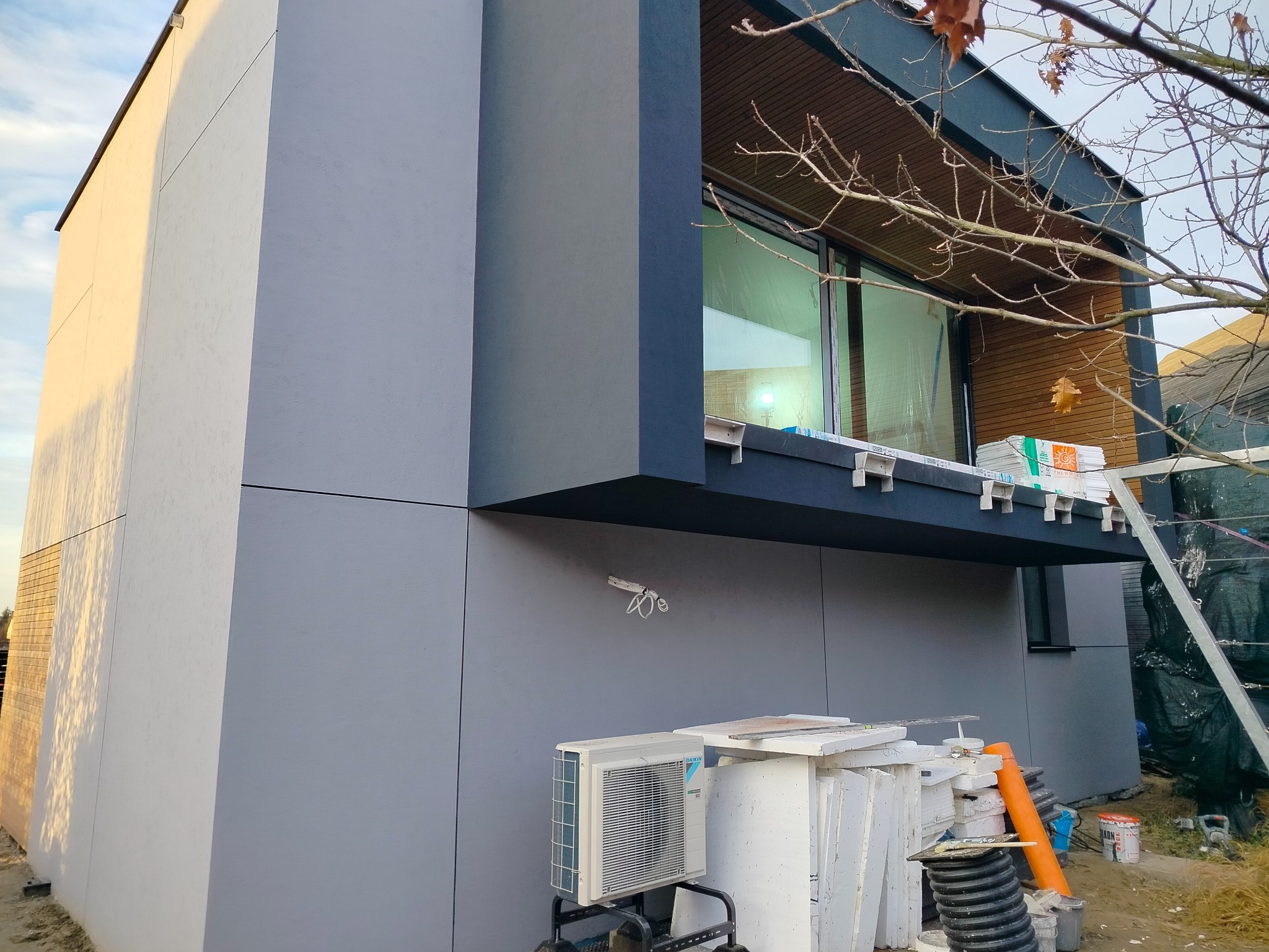 Nowoczesny dom z elewacją w odcieniach szarości, widoczne materiały budowlane i klimatyzator. Ujęcie z dołu, podkreślające geometrię budynku i detale wykończenia.