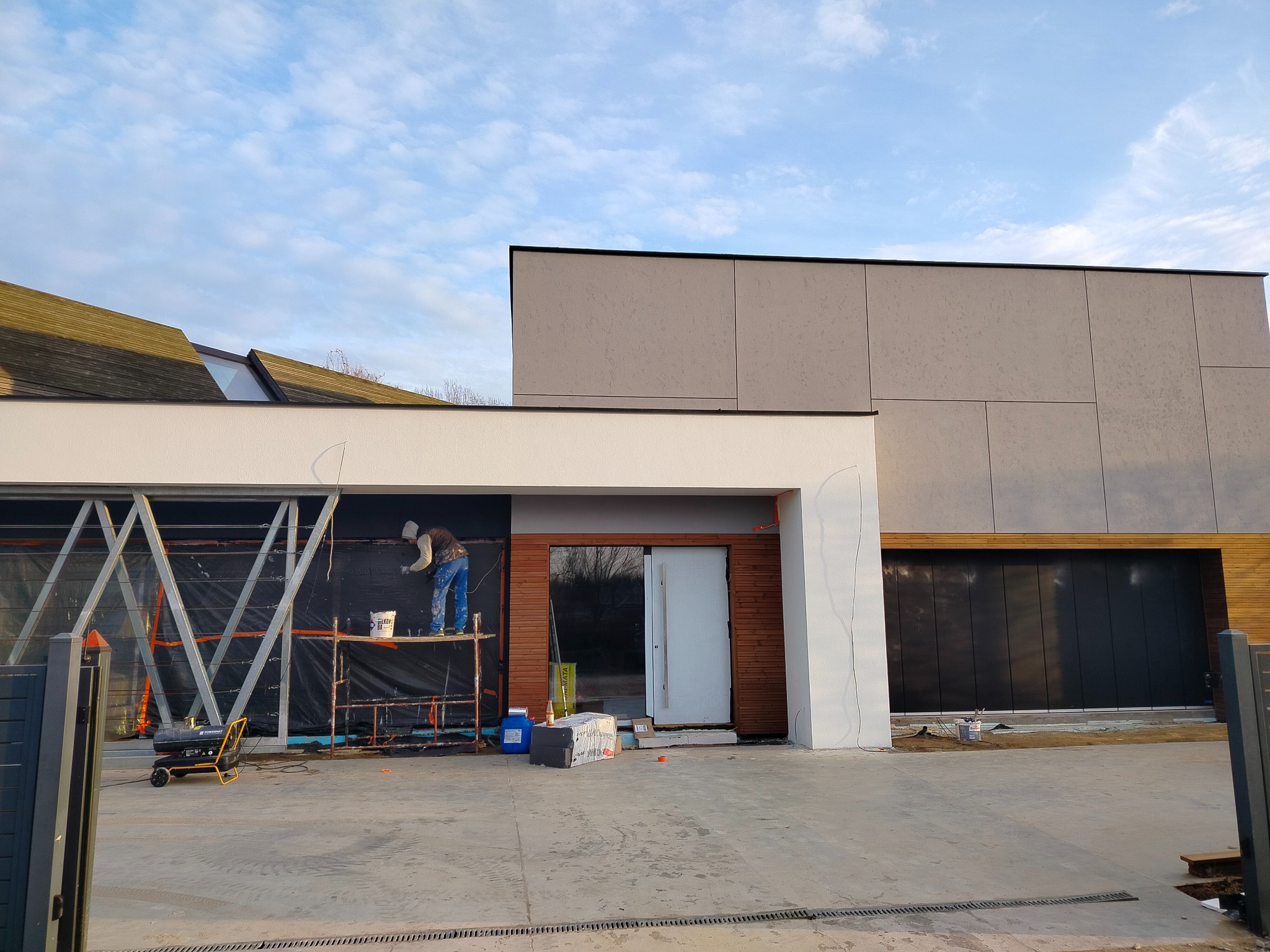 Pracownik na rusztowaniu maluje fragment elewacji nowoczesnego domu z elementami drewna i szarego tynku. Widoczne narzędzia i materiały budowlane.