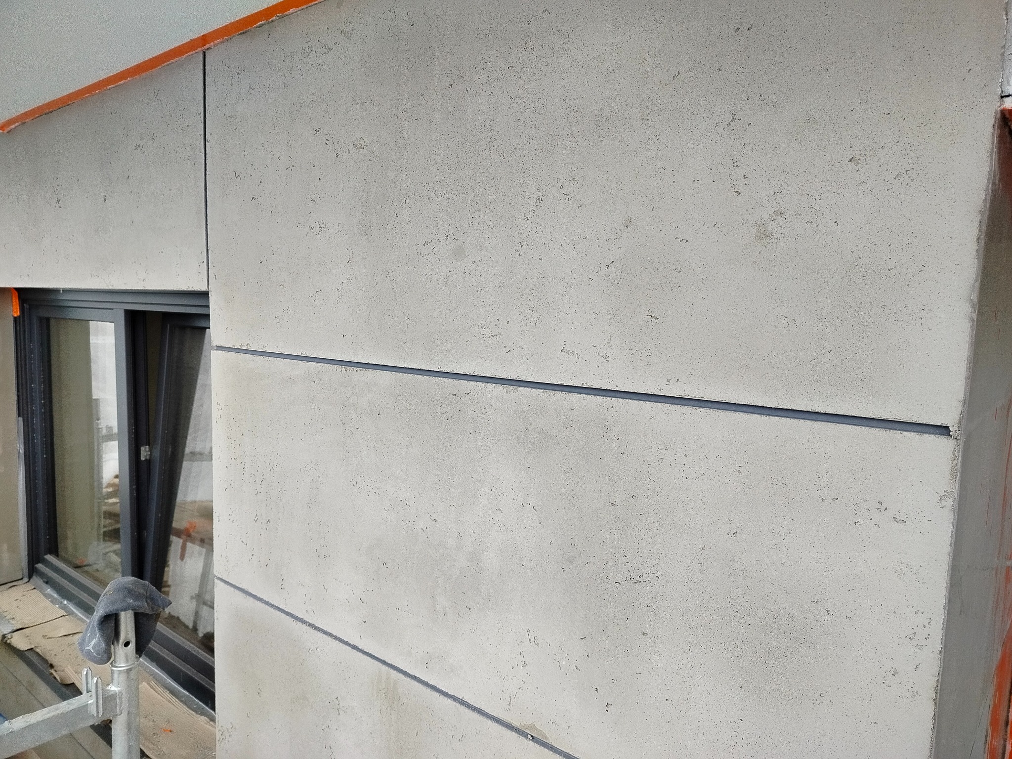 Szara elewacja budynku z widocznymi panelami i poziomymi szczelinami dylatacyjnymi. Fragment okna i rusztowania w tle. Detal wykończenia.